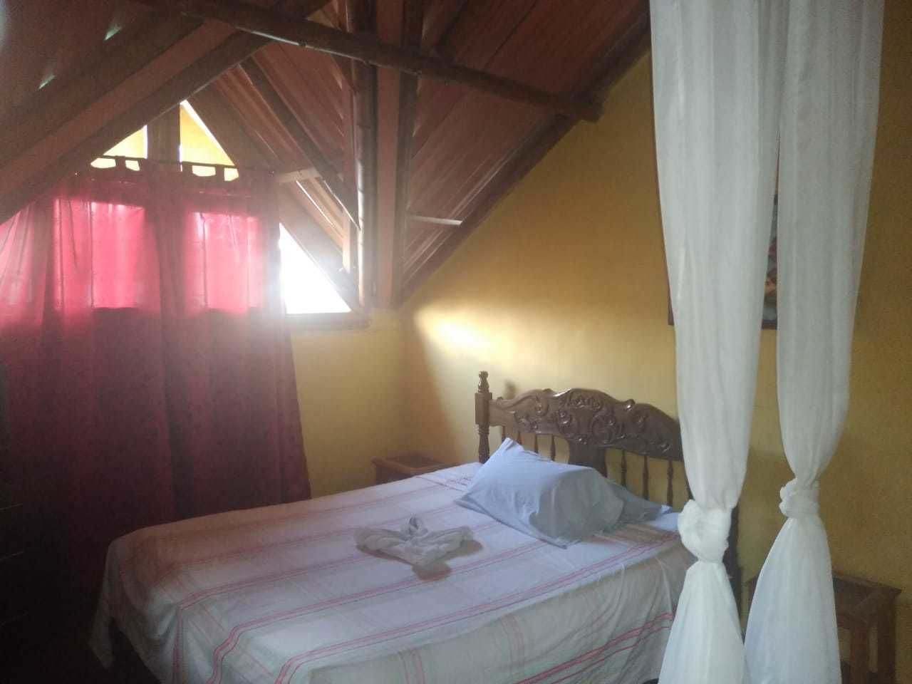Una cama individual con cabecera de madera en una habitación con techo inclinado y ventana con cortinas rojas.