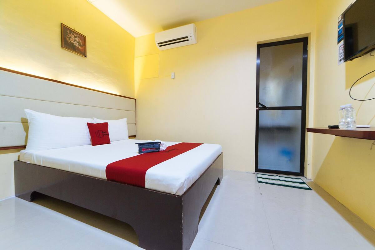 Une chambre d’hôtel avec un lit, un oreiller rouge, un couvre-lit blanc, une petite table de chevet, un téléviseur fixé au mur et un appareil de climatisation.
