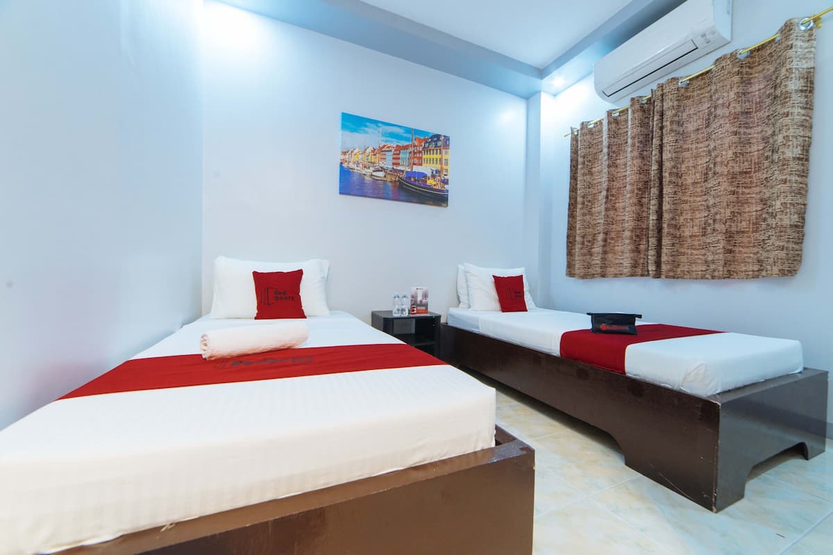 Une chambre d’hôtel avec deux lits, une couvre-lit rouge et blanche, un tableau représentant un paysage urbain et un rideau marron.