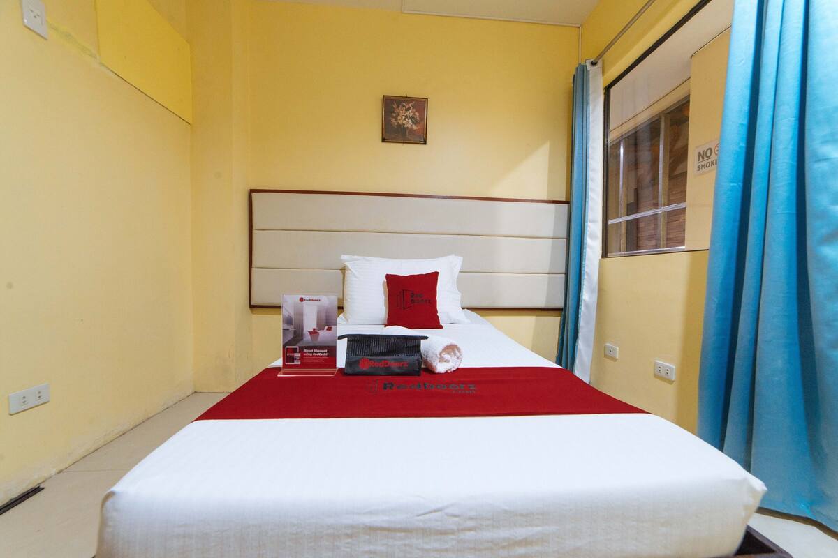 Une chambre d’hôtel avec un lit, une couverture rouge, un oreiller et une petite table sur laquelle se trouve un objet rouge et noir.