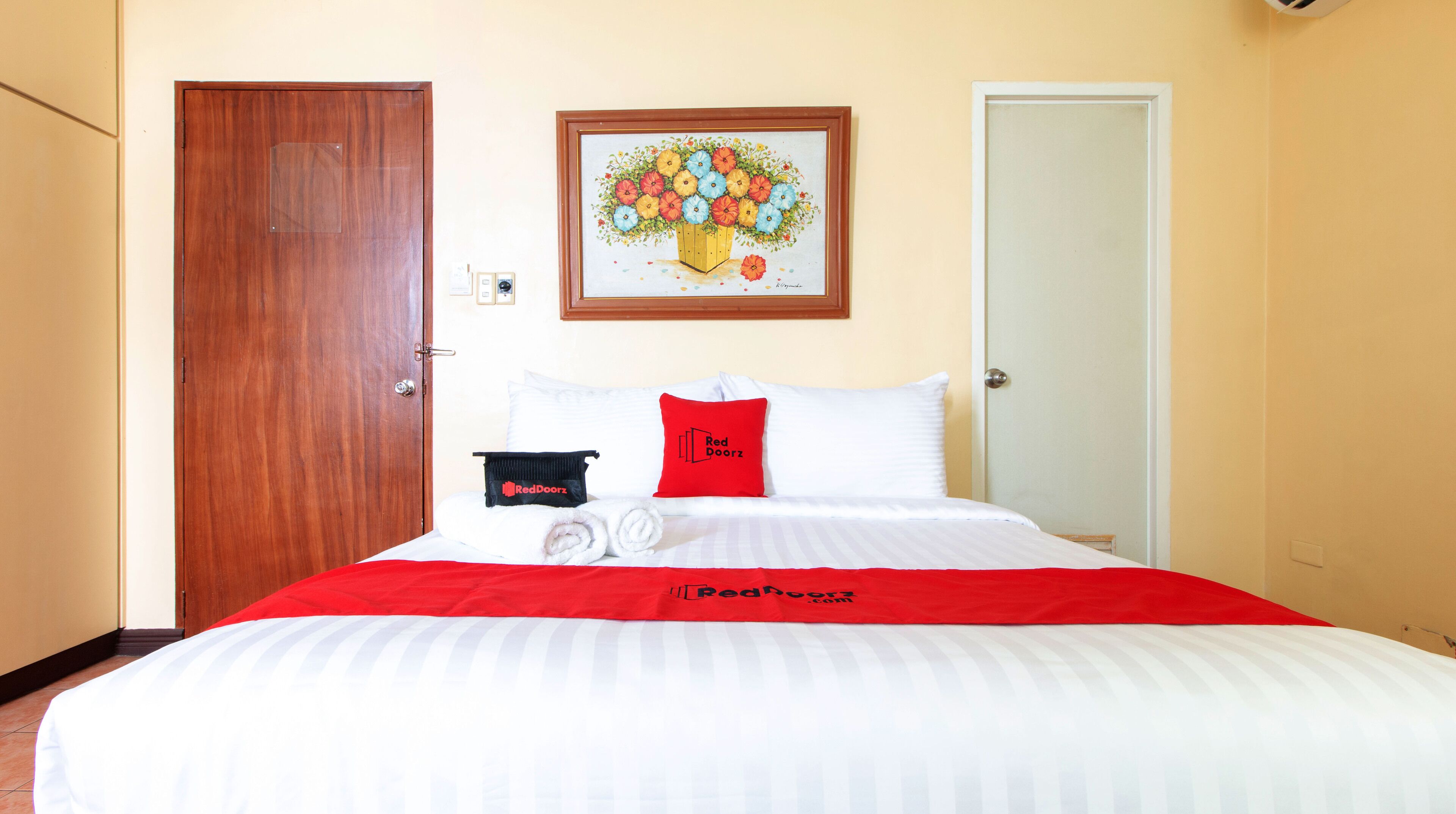 A hotel room with a bed, a red and white bedspread, a red pillow, a black towel, and a framed floral artwork on the wall.