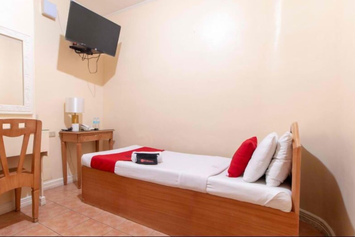 A small, single-bed room with a wooden headboard, a desk with a chair, a television mounted on the wall, and a bedside table with a lamp and a water bottle.