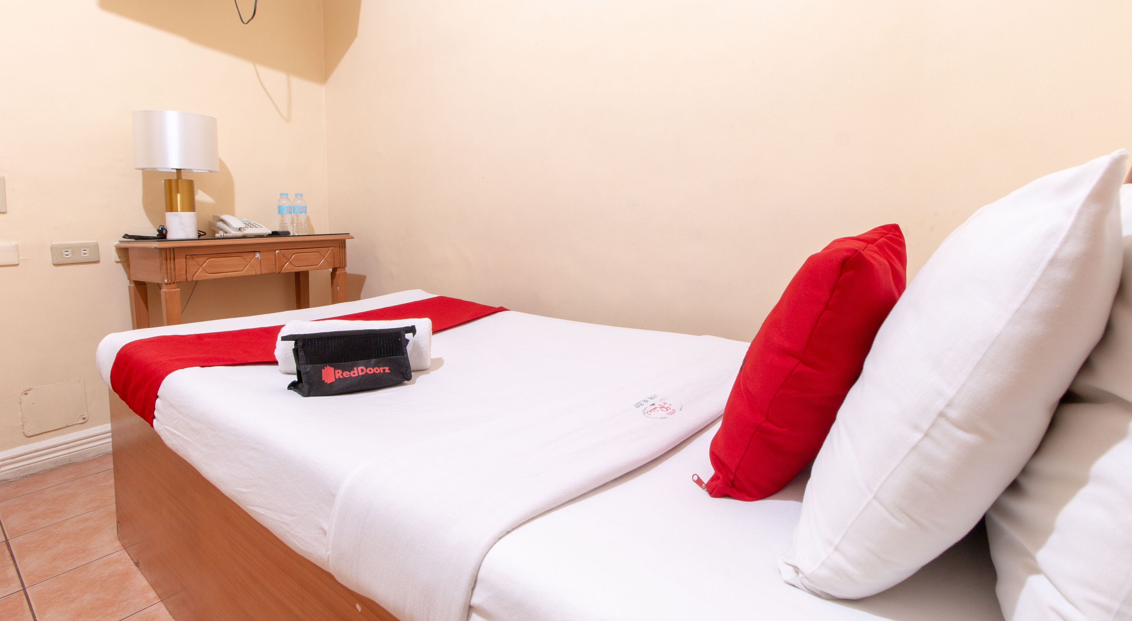 A neatly made bed with white and red bedding, a small bedside table with a lamp, a RedDoorz bag, and a wall socket.
