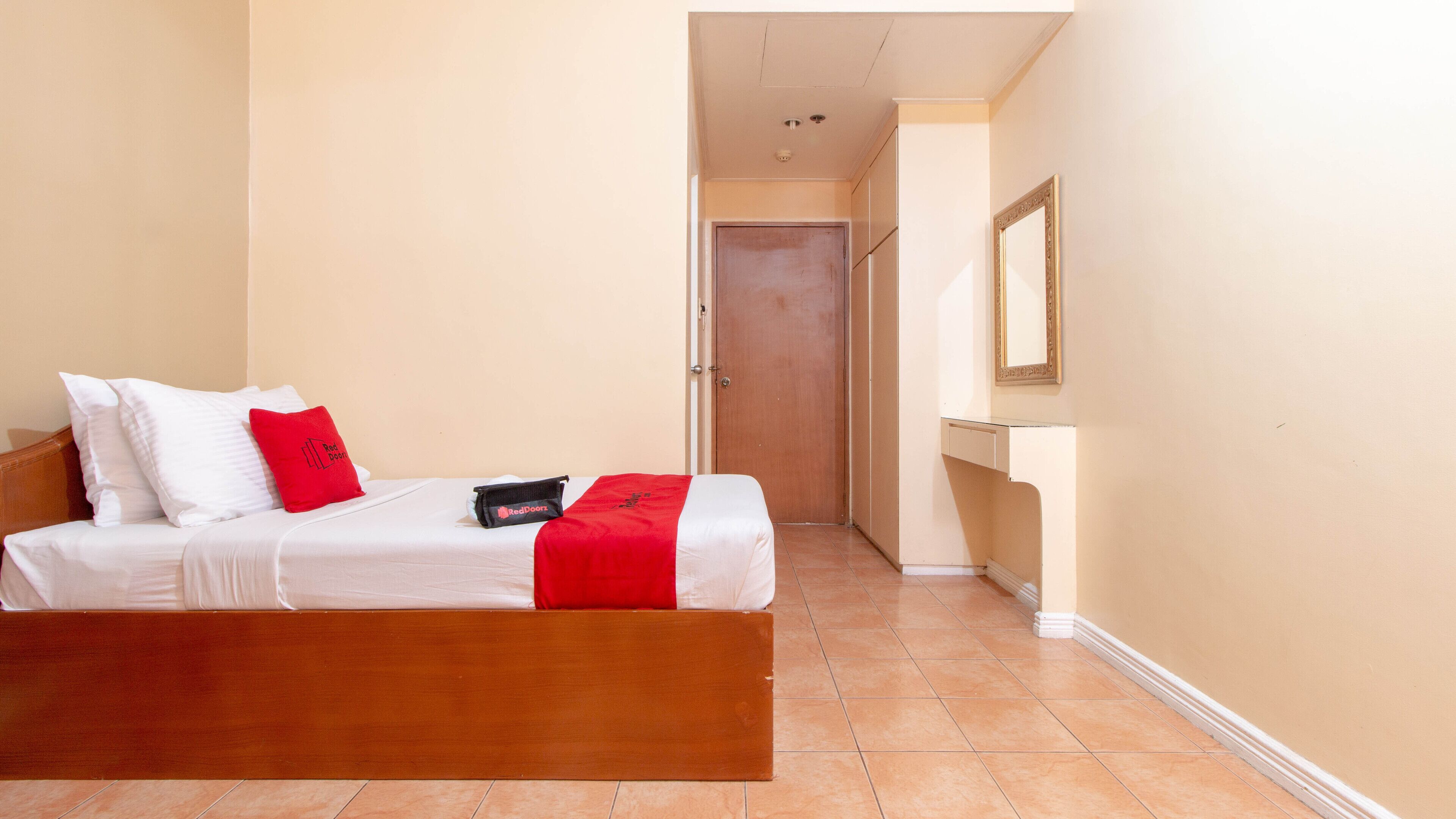 Ein Hotelzimmer mit einem hölzernen Bett, weißer Bettwäsche, einer roten Decke und einer RedDoorz-Tasche auf dem Bett.