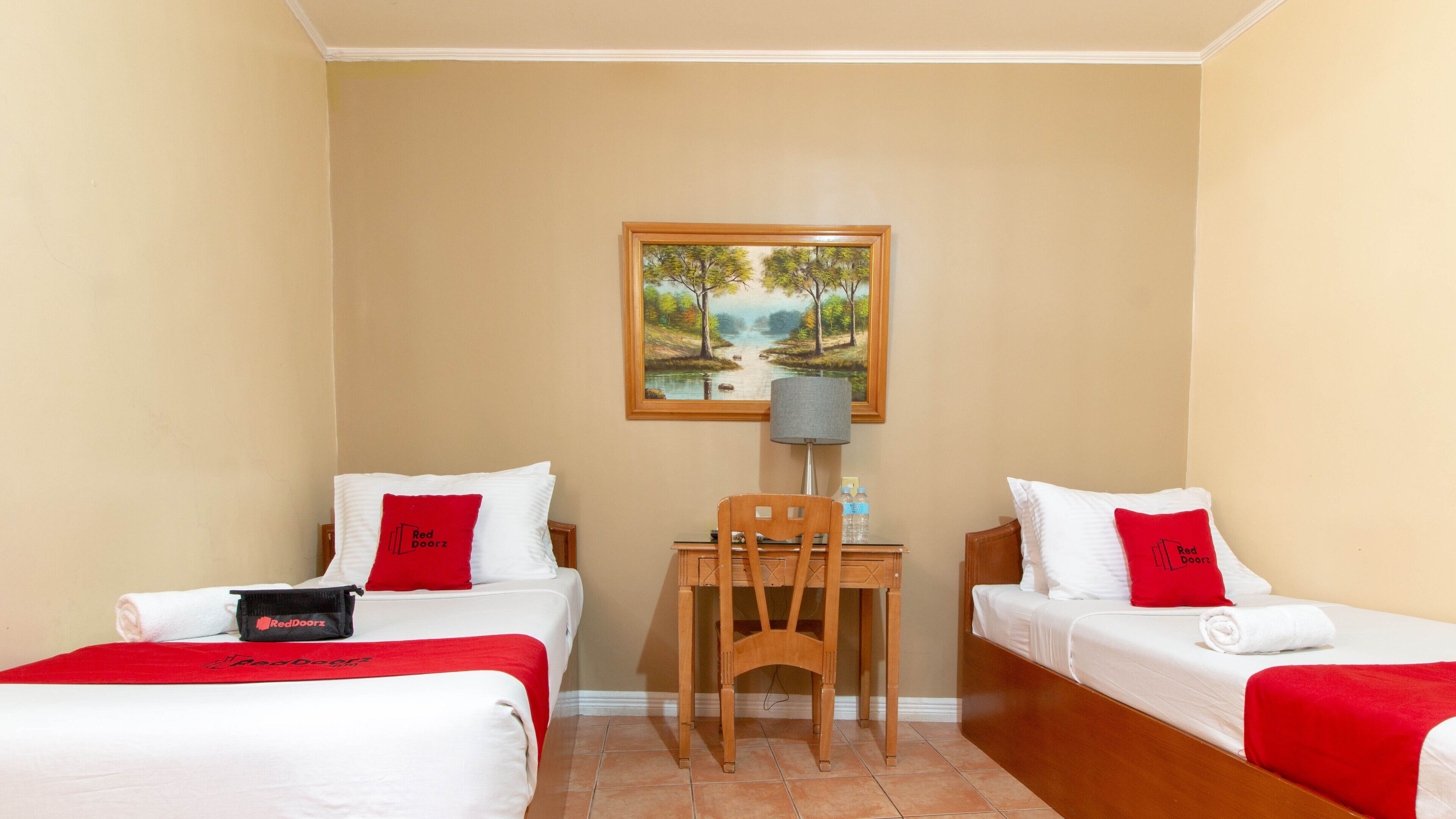 Habitación de hotel con dos camas individuales, un escritorio, una silla, una lámpara y un cuadro en la pared.