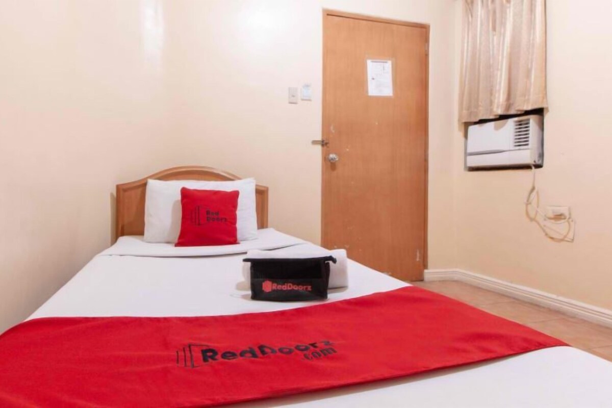 A single bed with a red blanket, a pillow, and a black bag on it.