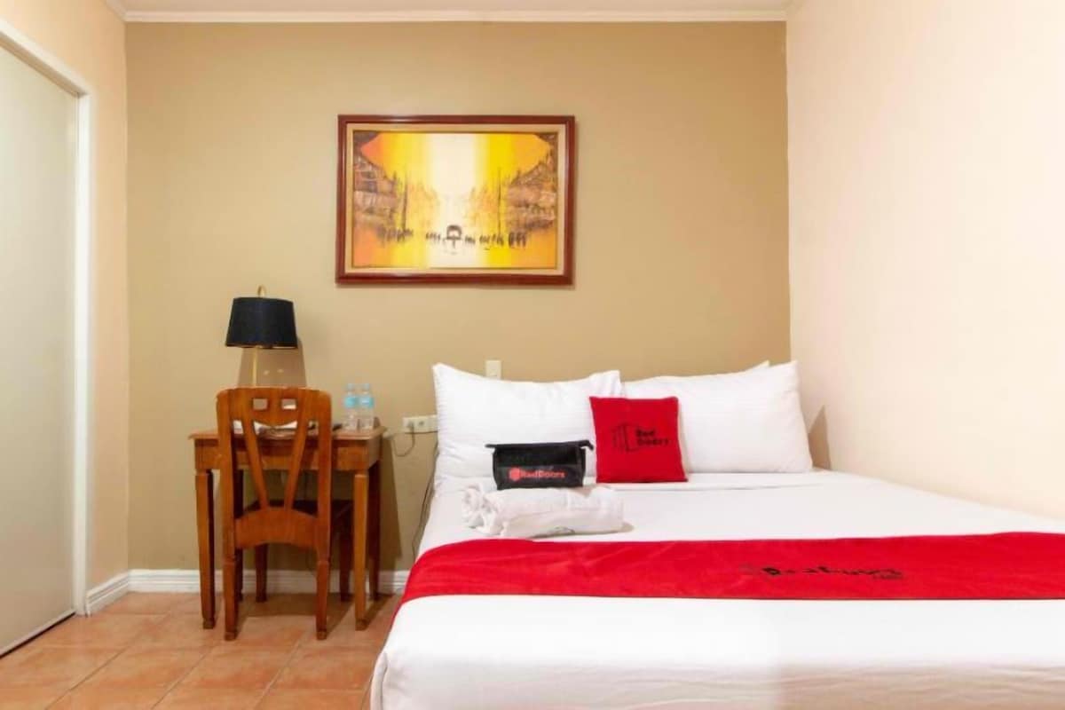 A hotel room with a bed, a desk, a chair, a lamp, and a painting on the wall.