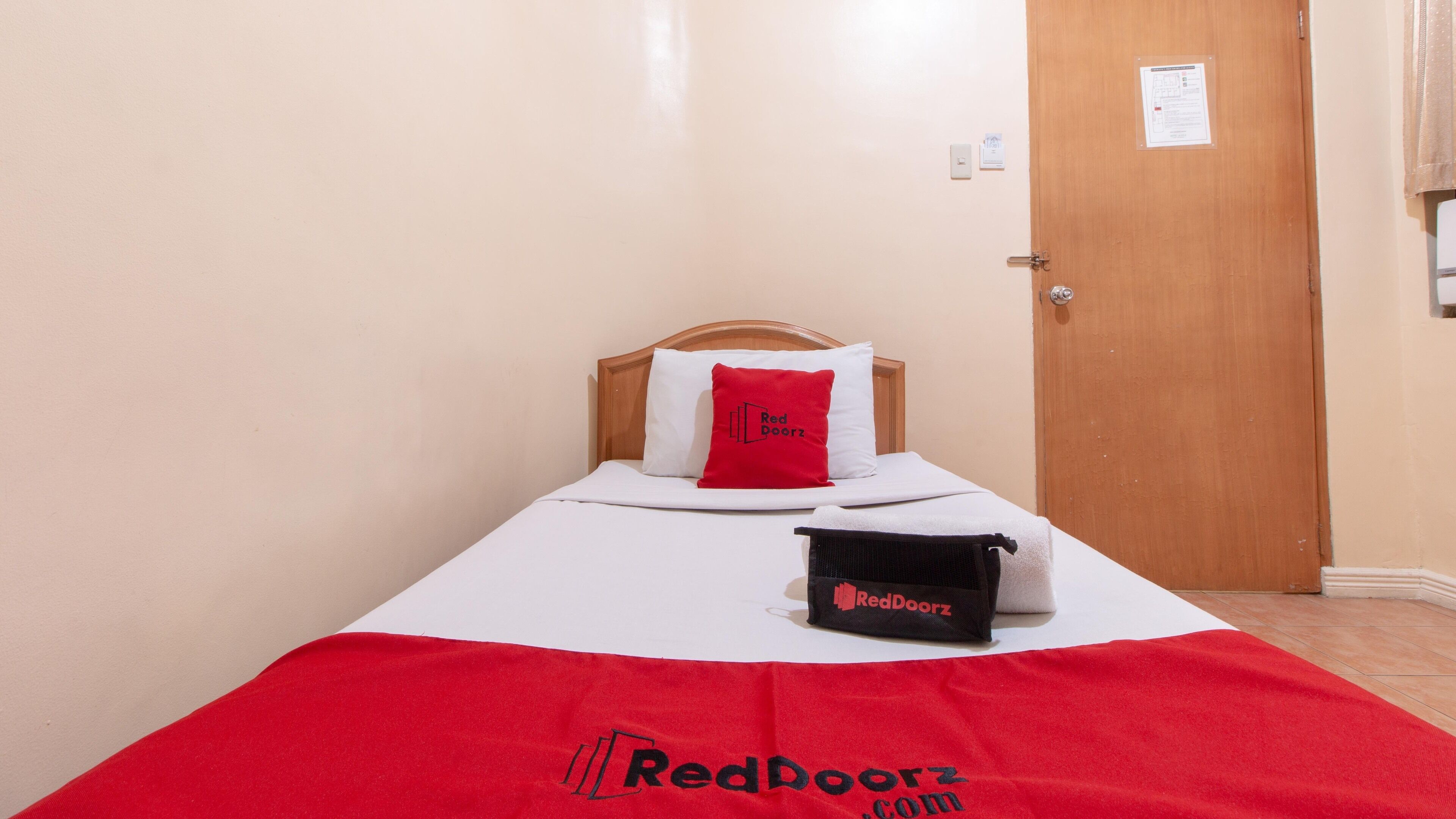 Una cama individual con colchón blanco y almohada roja, una bolsa negra con la etiqueta 'RedDoorz' y una puerta de madera en una habitación con paredes lisas.