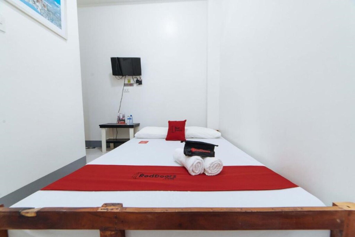 Une chambre d’hôtel avec un lit, une couverture rouge, un oreiller, un téléviseur fixé au mur et une petite table sur laquelle se trouvent une bouteille et un verre.