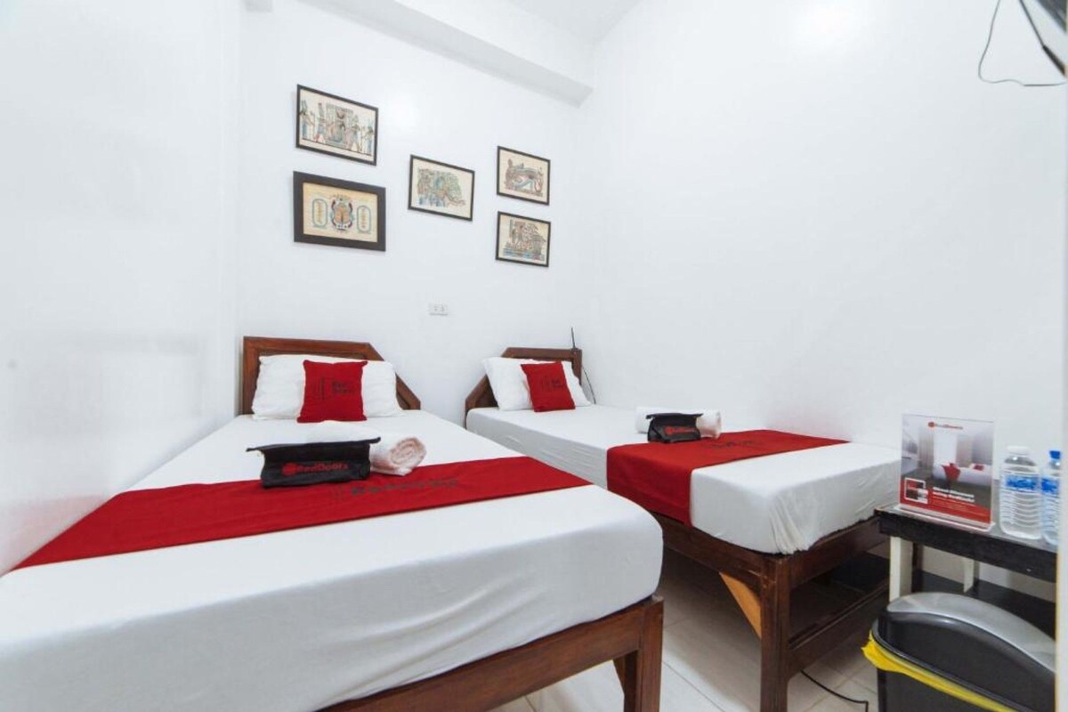 Une chambre d’hôtel avec deux lits simples, chacun recouvert d’une couverture rouge et blanche, et un petit bureau avec une chaise.