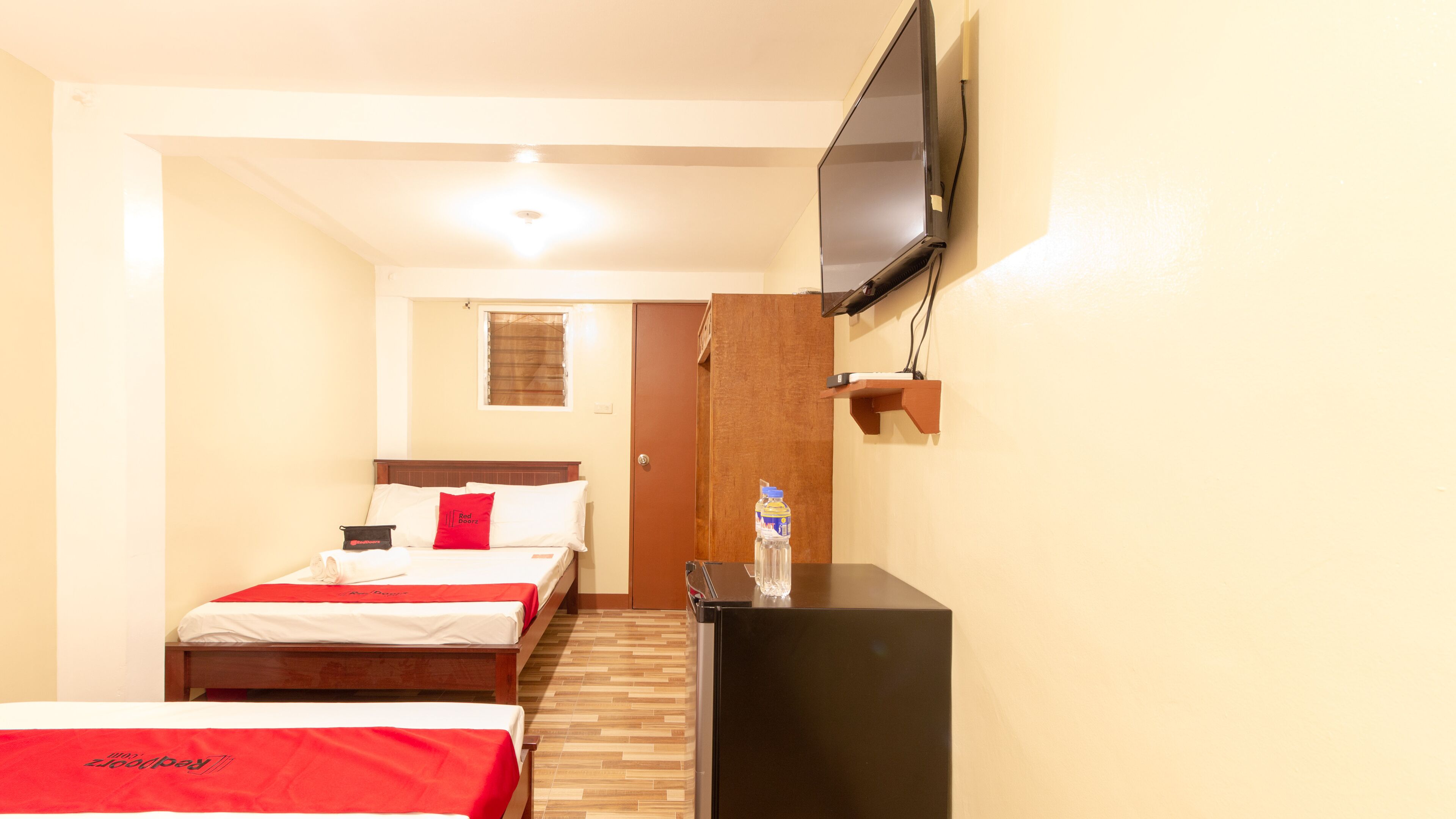 A hotel room with two beds, a TV mounted on the wall, a wooden floor, and a door leading to another room.
