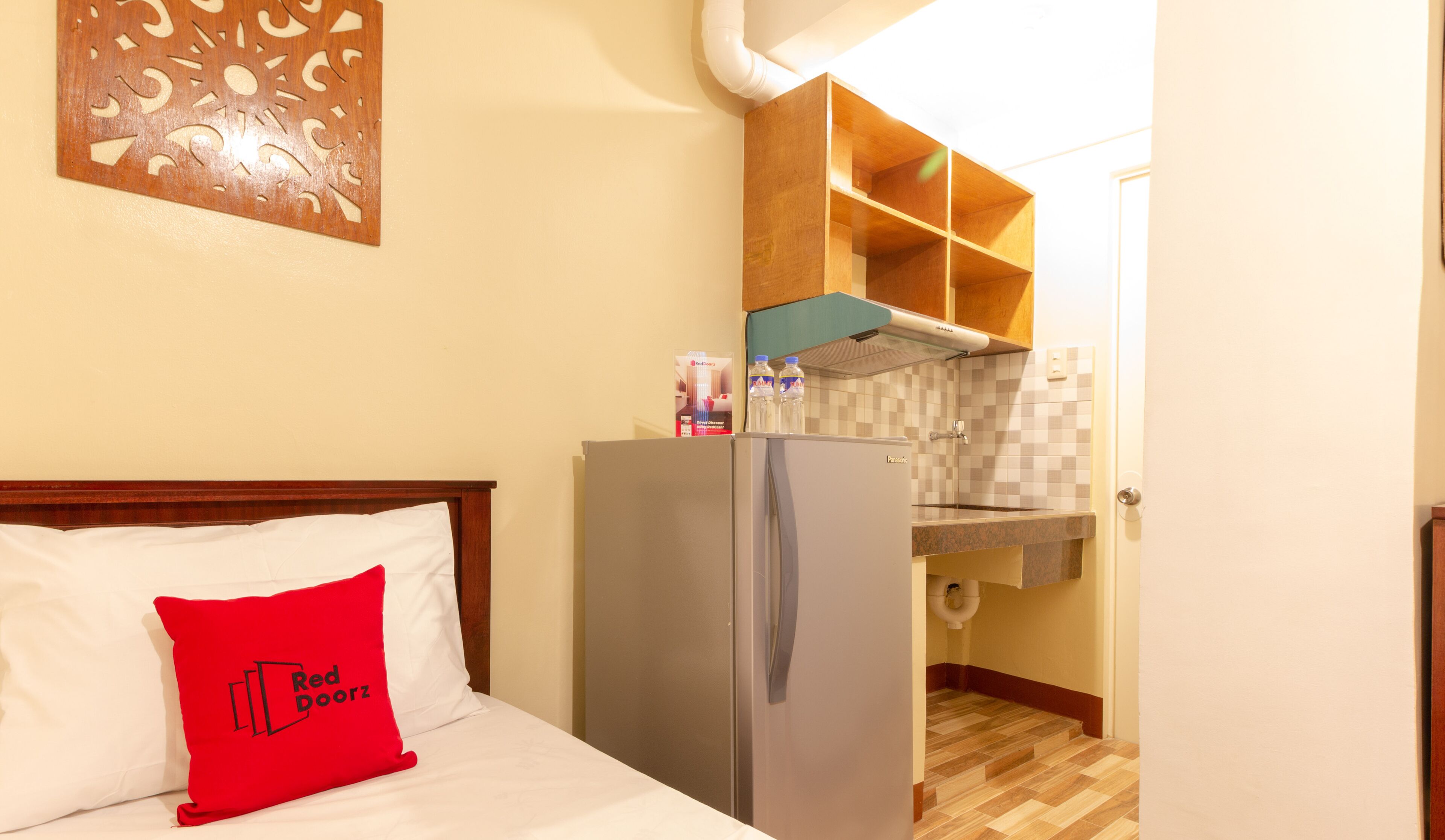 A compact hotel room with a bed, a red pillow, a refrigerator, a kitchenette, and a wooden wall decoration.