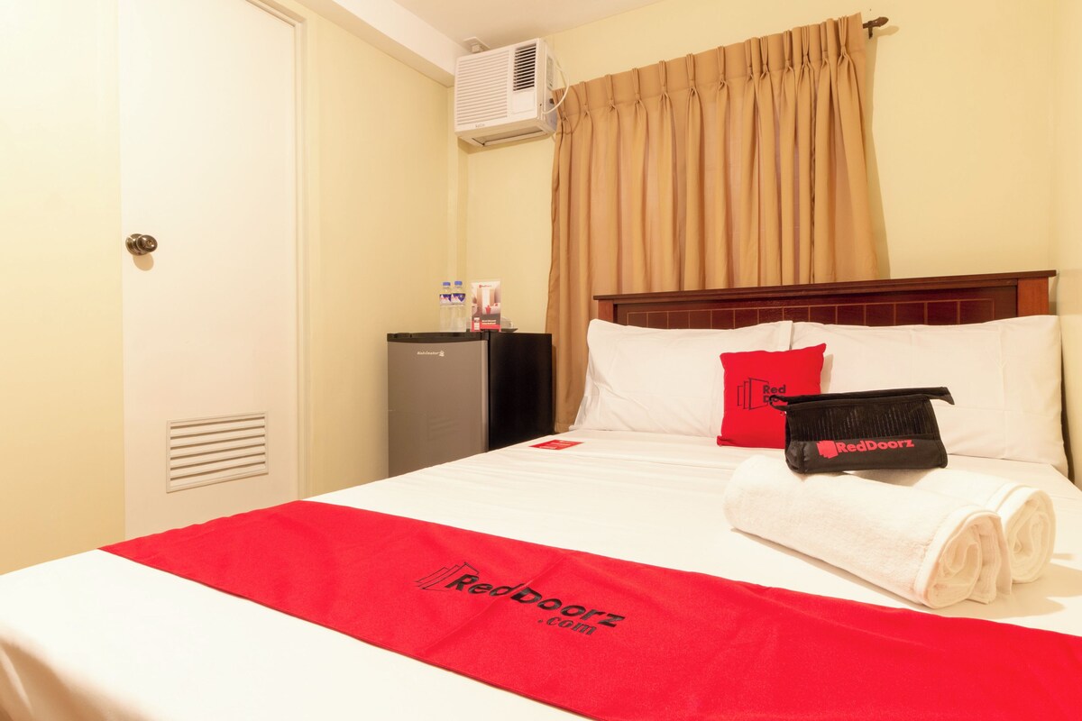 Ein Hotelzimmer mit einem Bett, einer rot-weißen Tagesdecke, einem roten Kopfkissen, einer schwarzen Tasche, einer Minibar und einem Fenster mit beigen Vorhängen.
