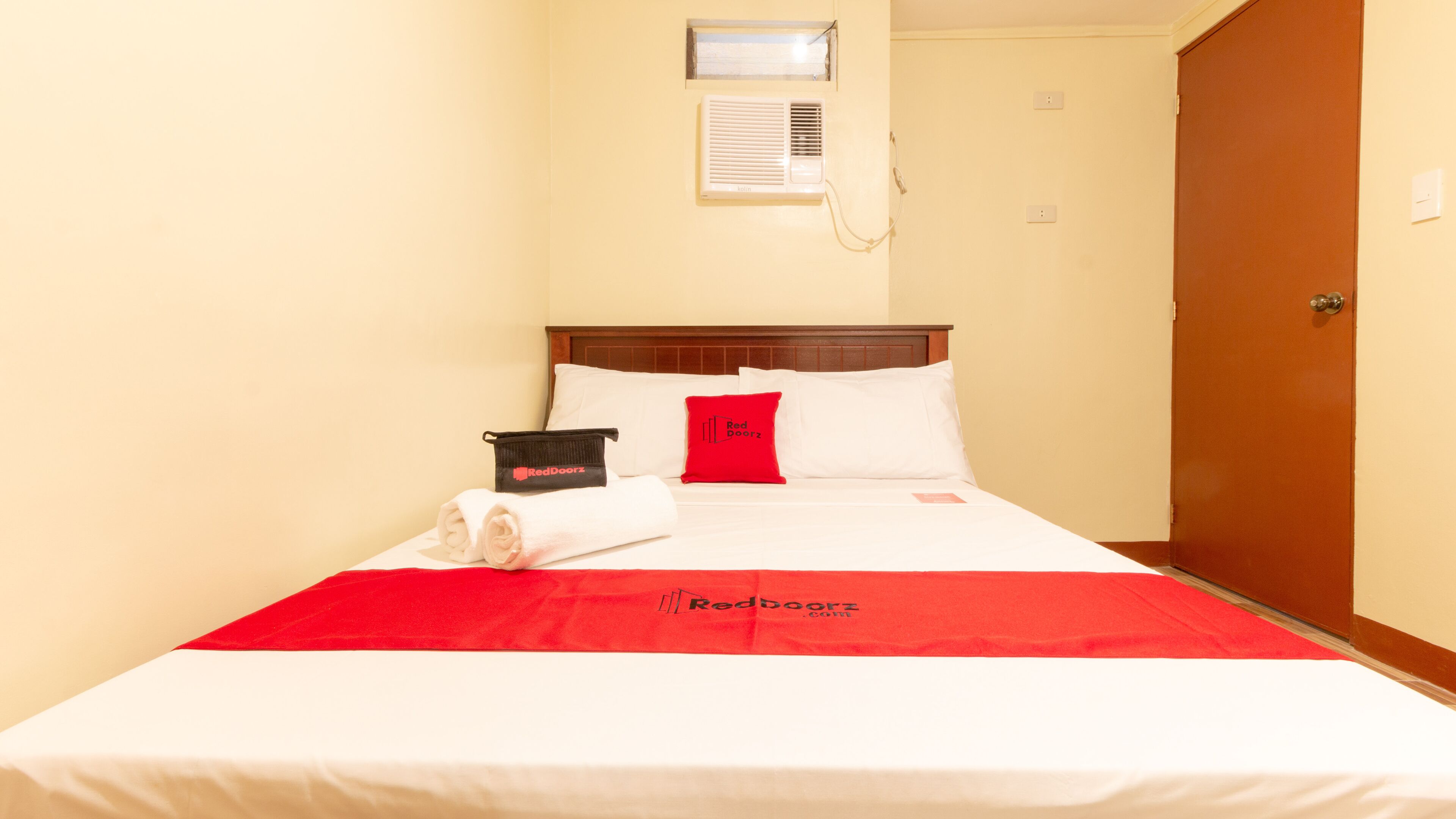 A hotel room with a bed, a red and white bedspread, a red pillow, a black bag, and a wall-mounted air conditioning unit.