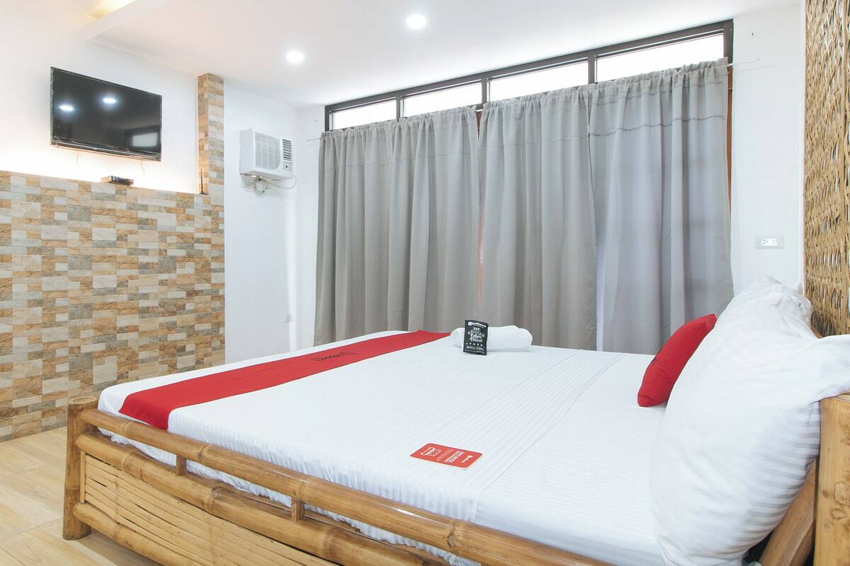 A hotel room with a bed, a TV mounted on the wall, a window with curtains, and a stone-patterned wall.