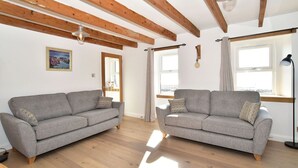 Un salón espacioso con dos sofás grises, vigas de madera en el techo y ventanas grandes que dejan entrar luz natural.