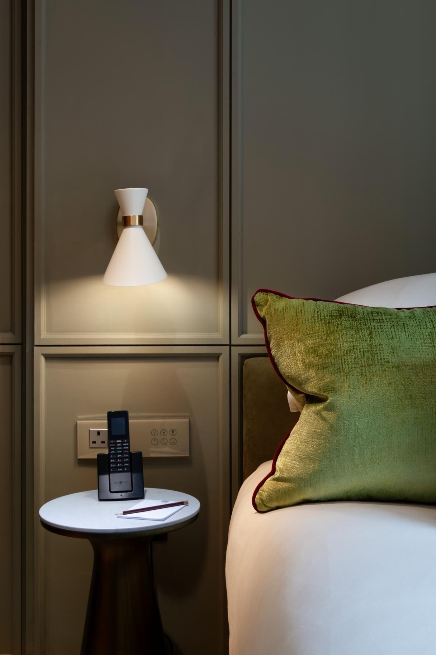 Een hotelkamer met een bed, een nachtkastje met een telefoon, een wandlamp en een groen kussen.