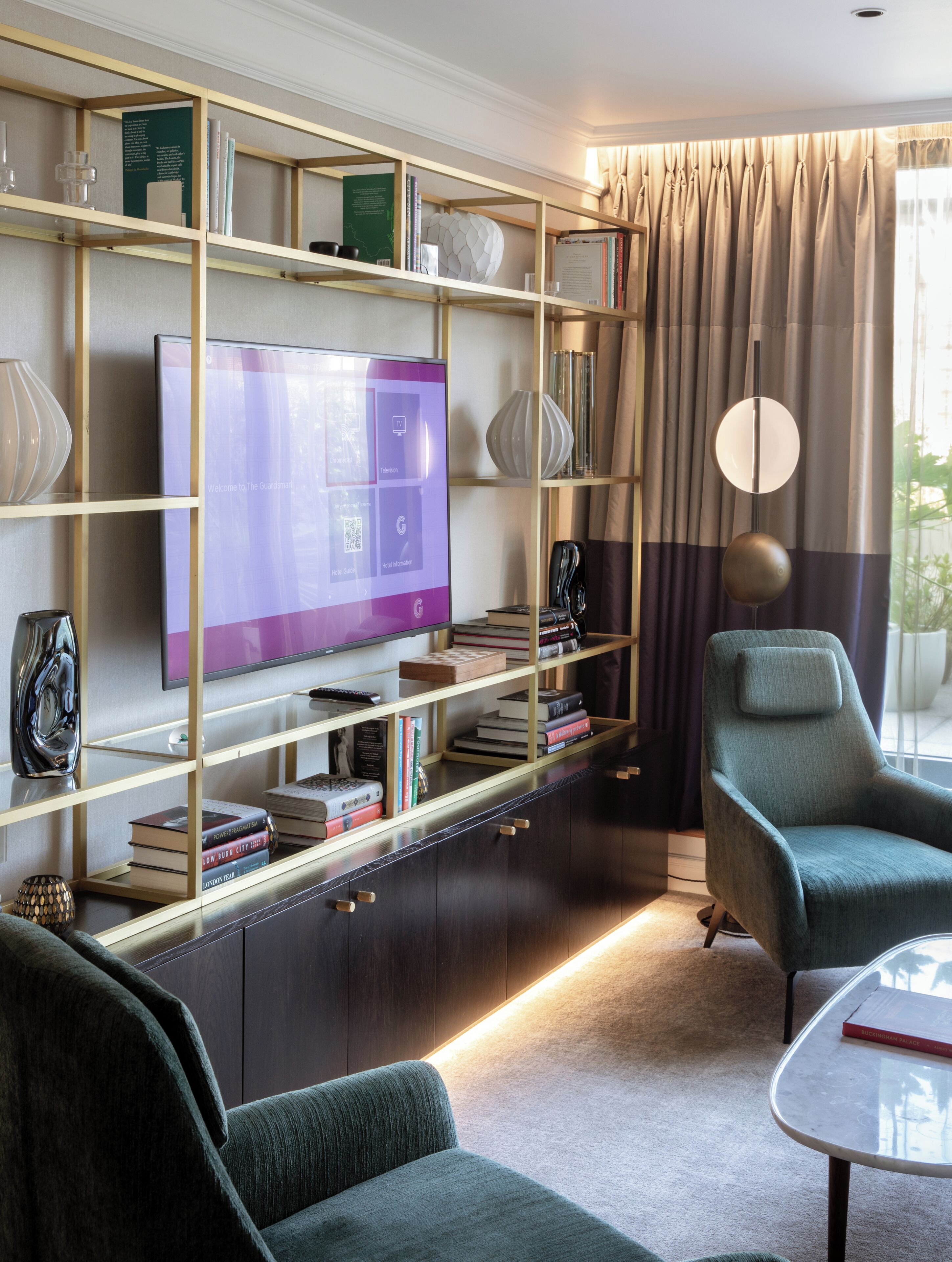 A modern hotel room with a large flat-screen TV, a bookshelf with books and decorative items, a comfortable armchair, and a small table.