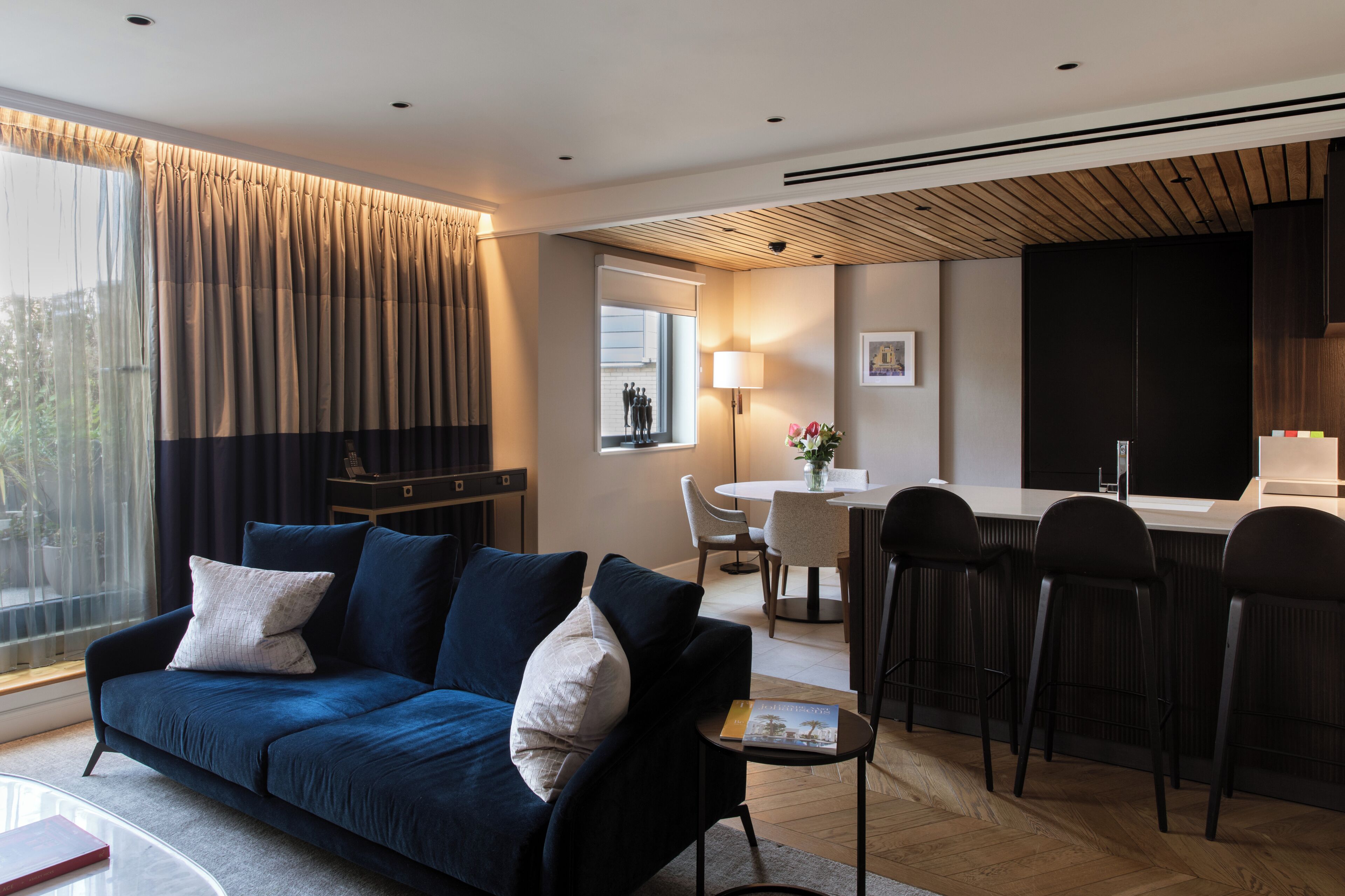A modern living room with a dark blue sofa, a dining area with a round table, and a kitchen with a dark cabinet.
