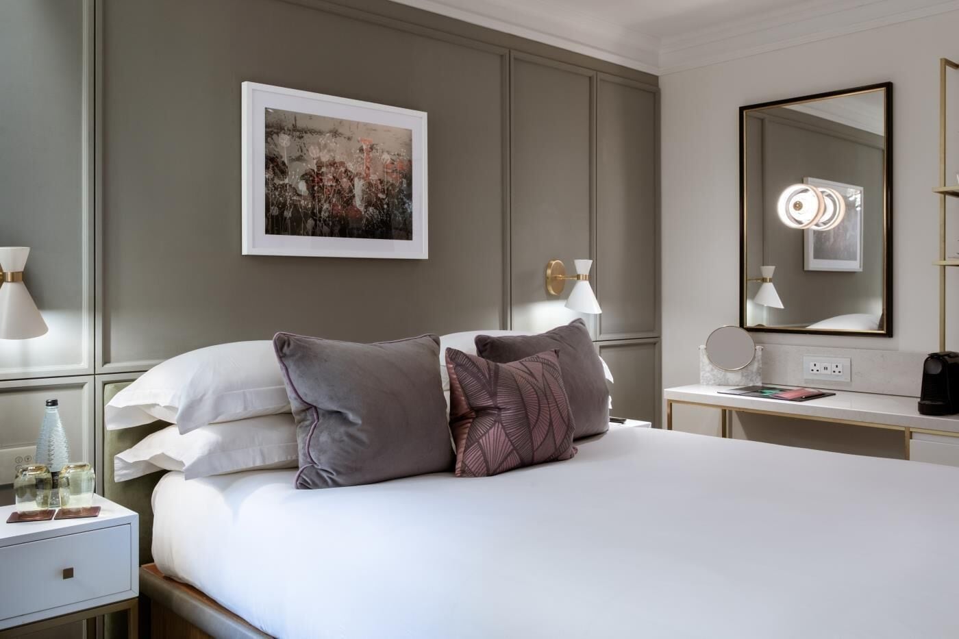 A hotel room with a bed, bedside tables, a desk, a mirror, and a framed artwork on the wall.