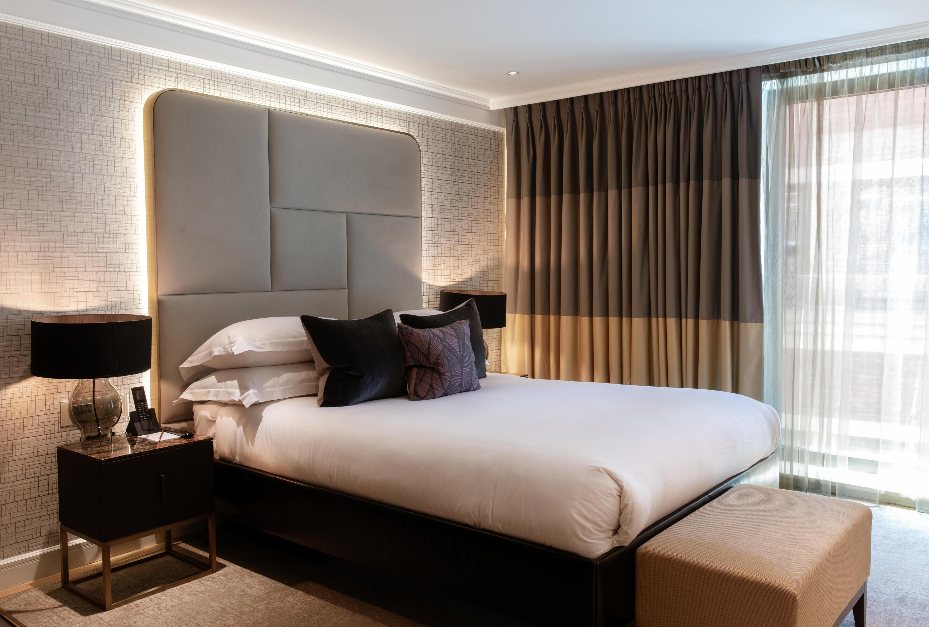 A hotel room with a large bed, a headboard, a nightstand with a lamp, and a window with curtains.