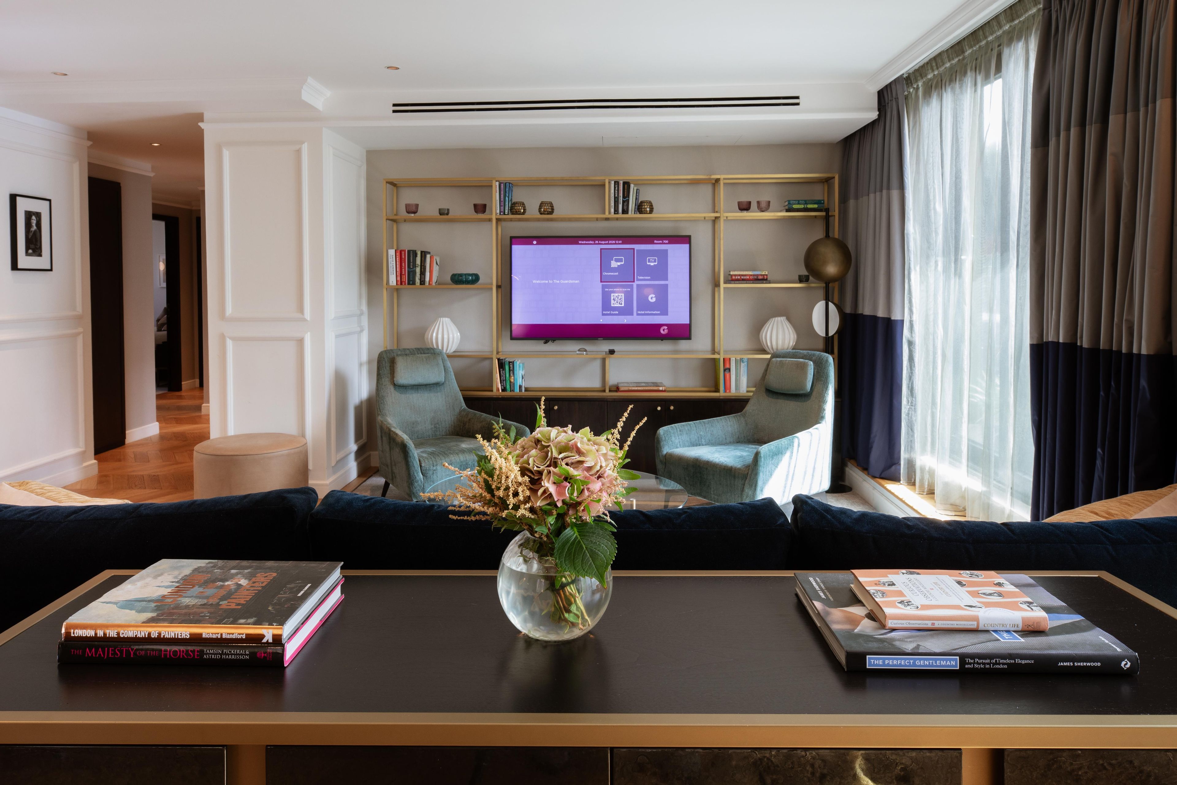 A modern living room with a flat-screen TV, two armchairs, a coffee table with books, and a vase with flowers.