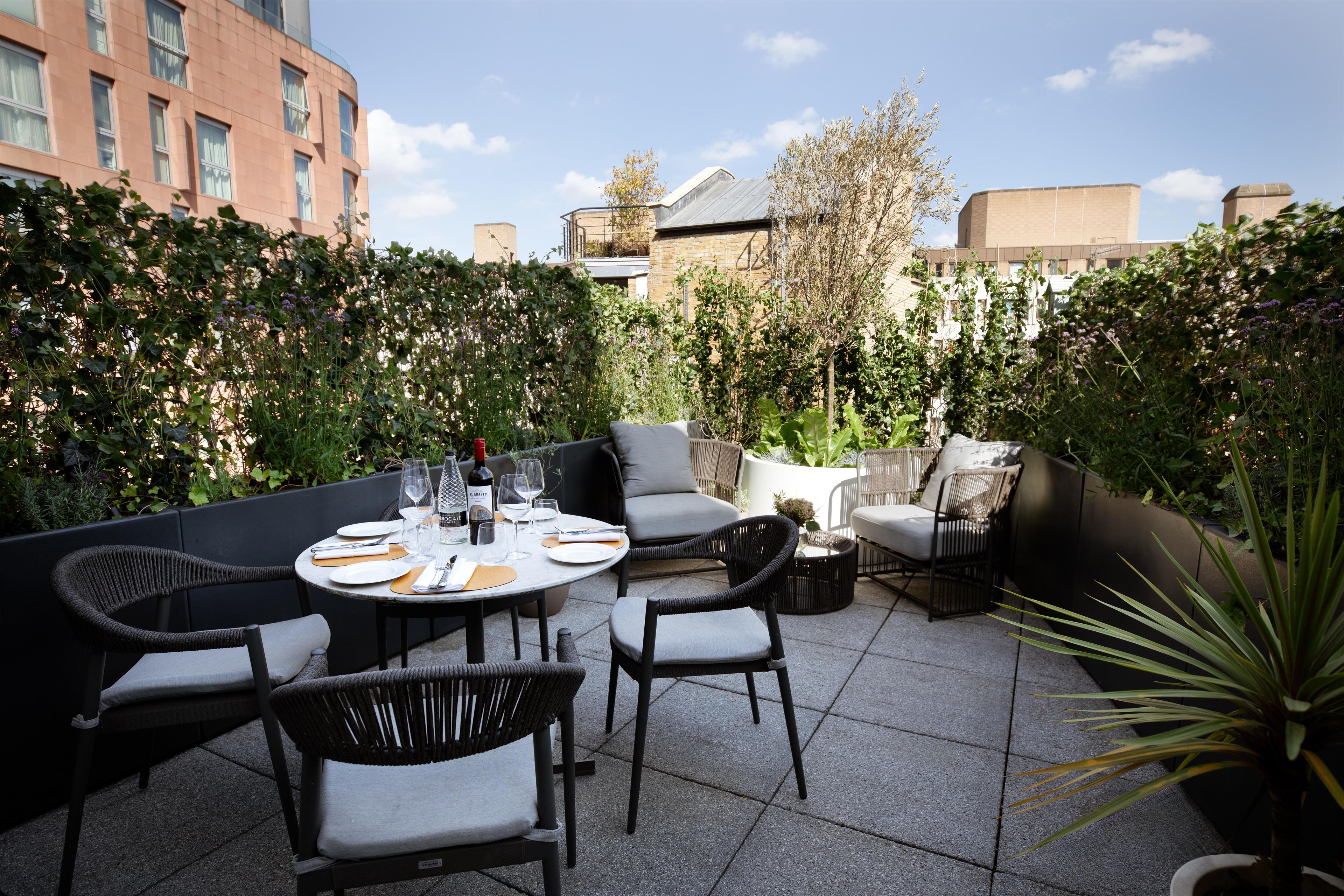 Une terrasse extérieure avec une table ronde dressée pour le repas, entourée de chaises en osier et d’un coin salon agrémenté de coussins.