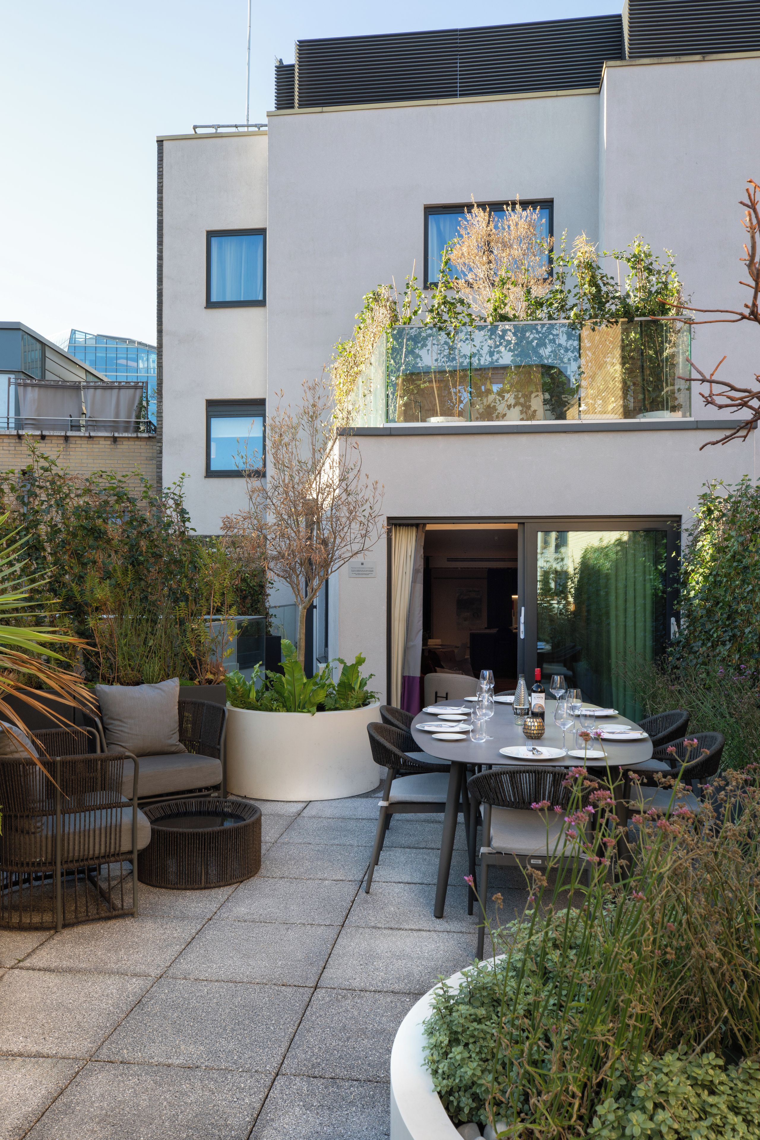A modern outdoor dining area with a table set for two, surrounded by wicker furniture and potted plants.