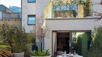 A modern outdoor dining area with a table set for two, surrounded by wicker furniture and potted plants.