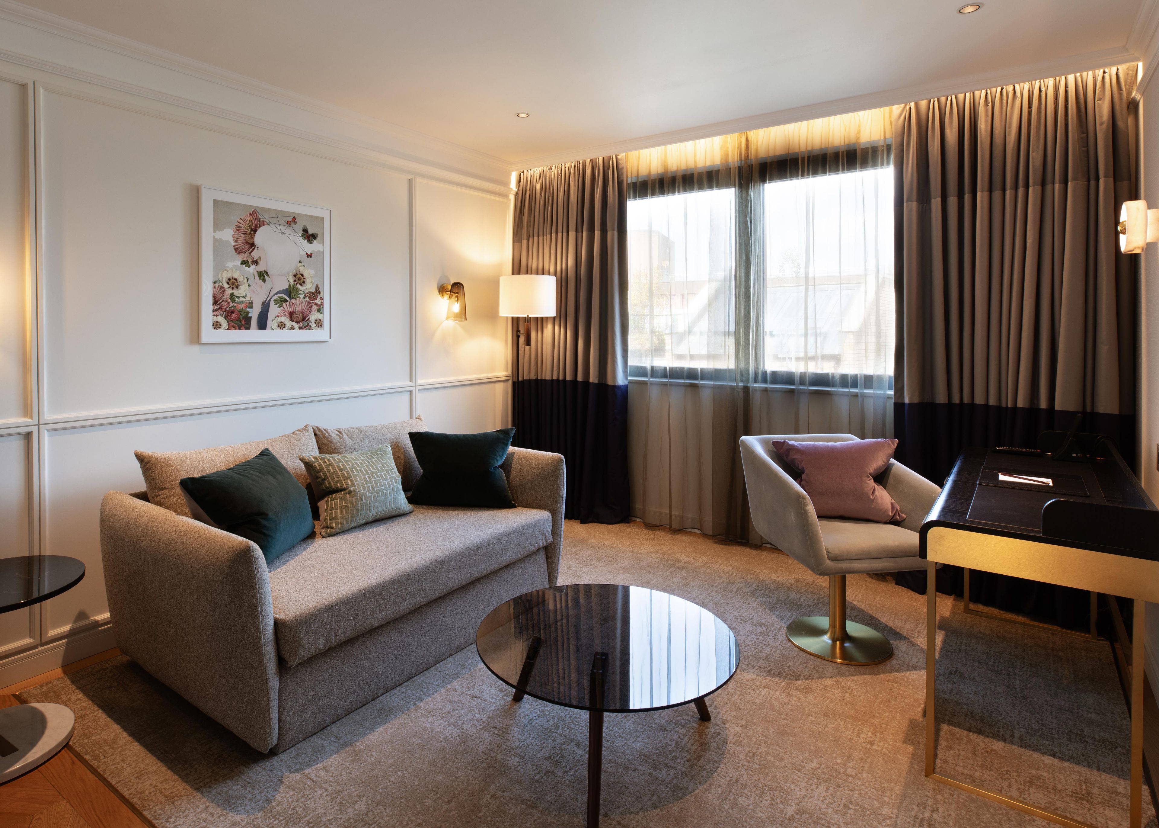 Una habitación de hotel moderna con sofá, sillón, mesita redonda y escritorio con lámpara.