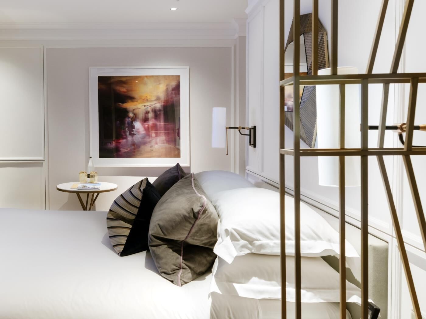 A hotel room with a bed, pillows, a small table with a bottle and a book, a painting on the wall, and a window with a metal grille.