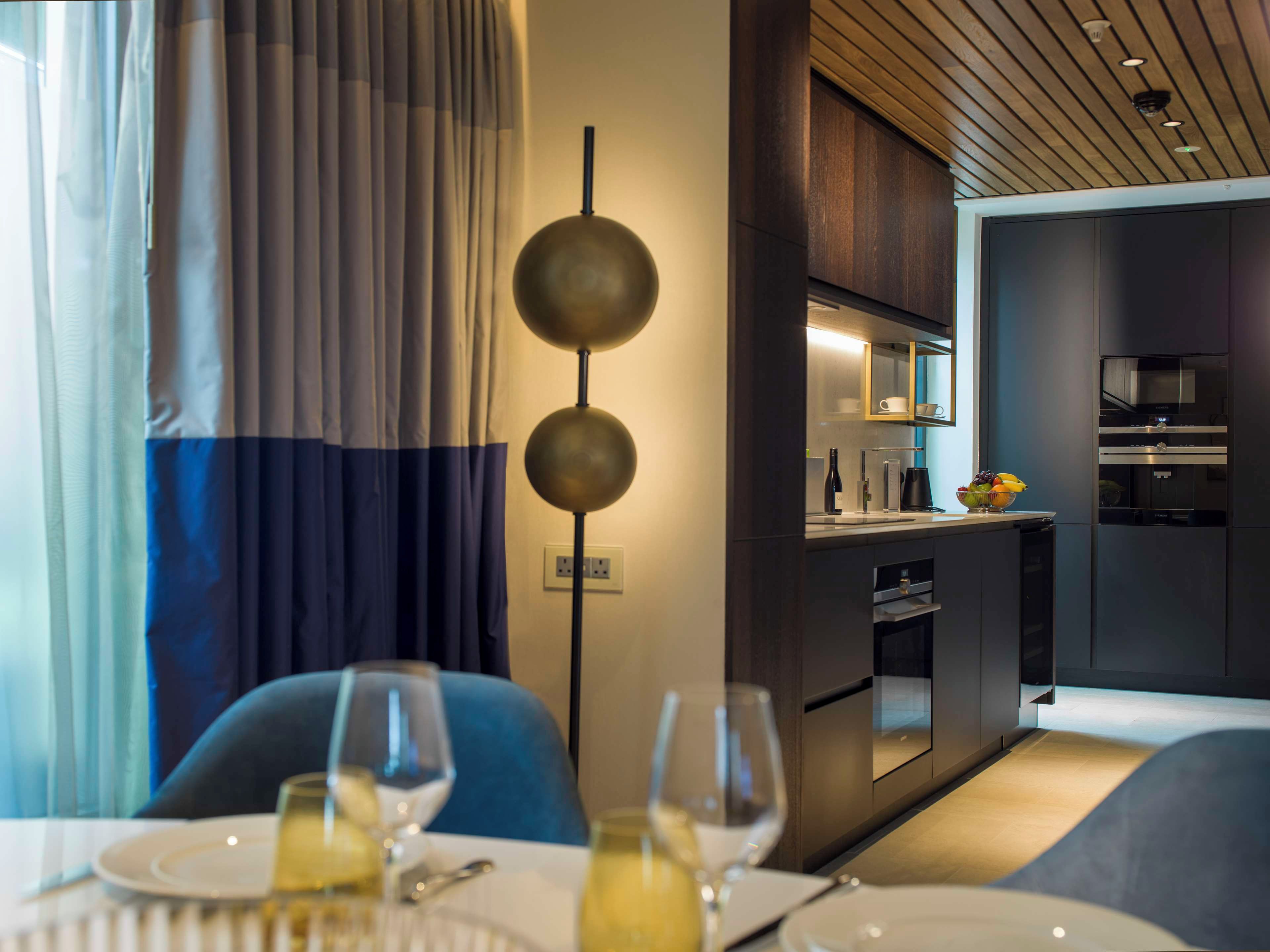 Un espace repas moderne avec une table dressée pour deux personnes, une cuisine en arrière-plan et un lampadaire doté de deux abat-jour sphériques.