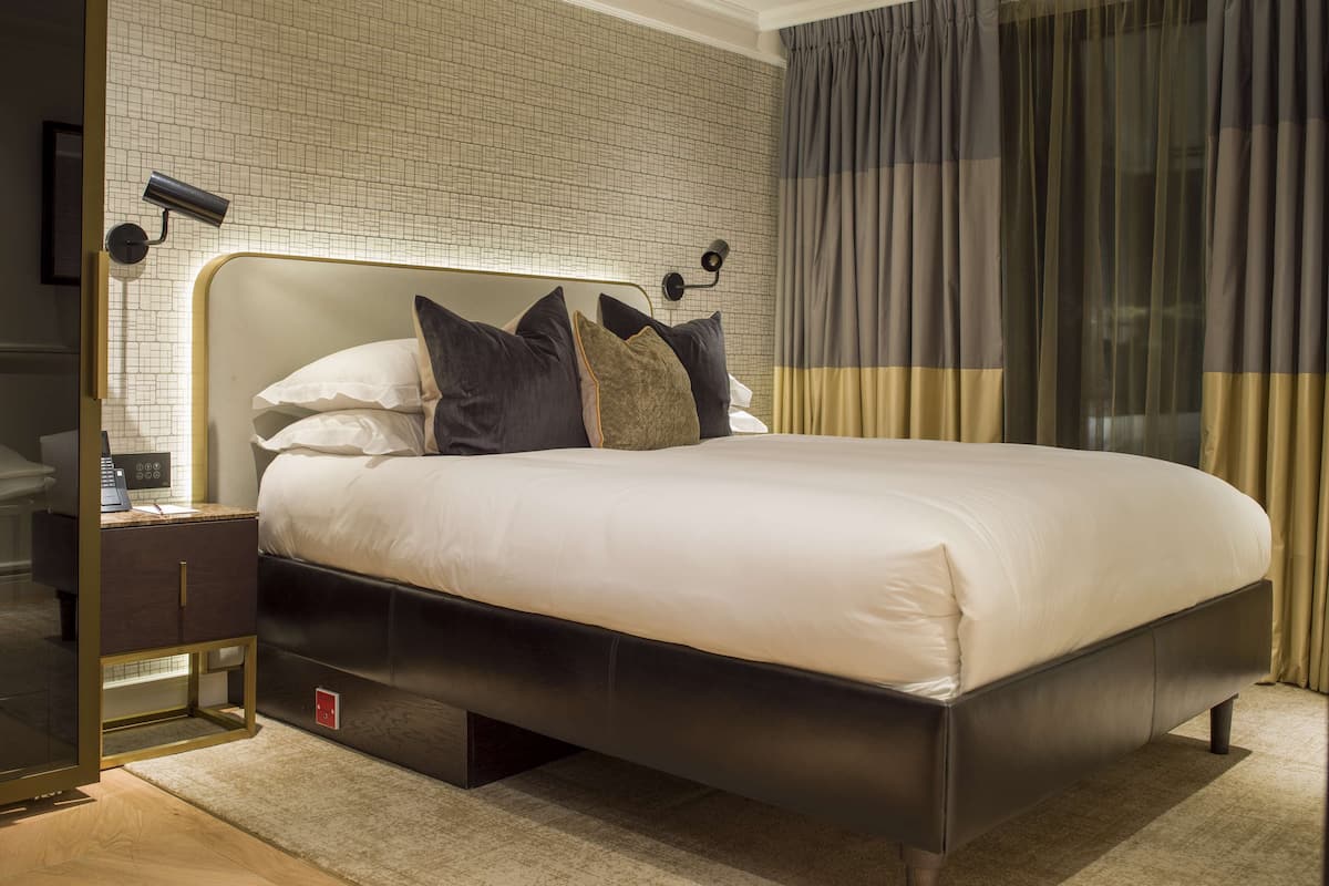 A modern hotel room with a large bed, a nightstand, a wall-mounted lamp, and a view of the city through sheer curtains.
