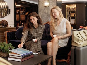 Two individuals seated on a sofa with a stack of books and a handbag on a table. - The Guardsman Hotel and Residences (London)