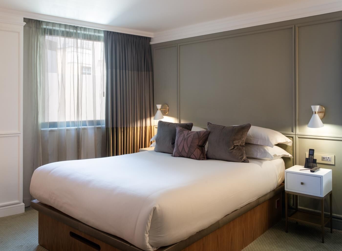 A hotel room with a large bed, a bedside table with a lamp, and a view of the outside through a window with curtains.