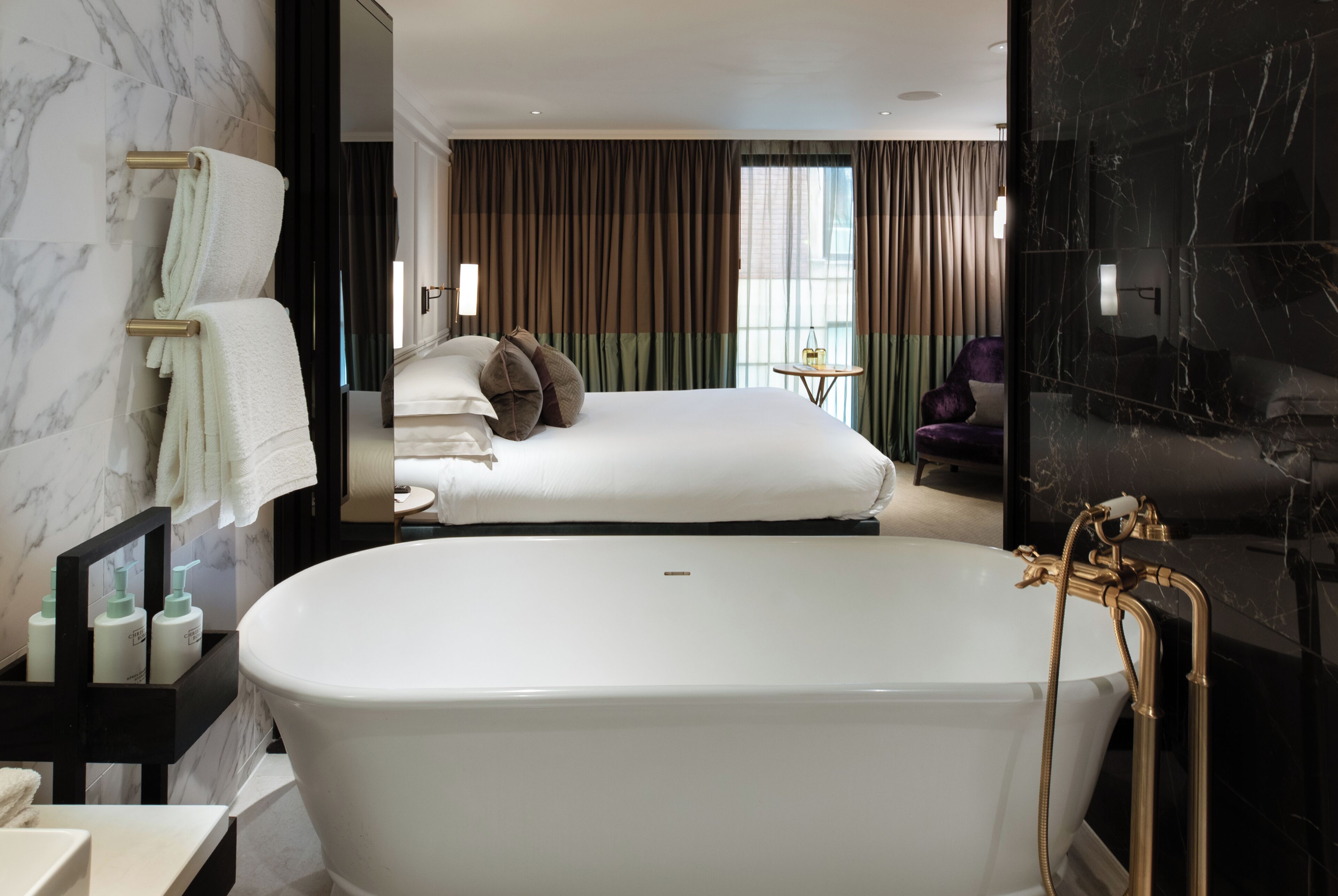 A modern hotel room with a large bathtub, a bed with multiple pillows, a small round table, and a chair.