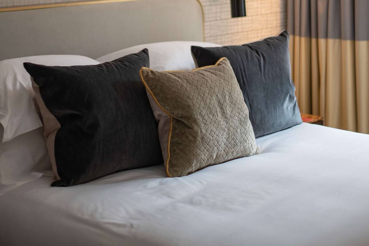 Cama bem arrumada com lençóis brancos e três almofadas em preto, bege e cinza.