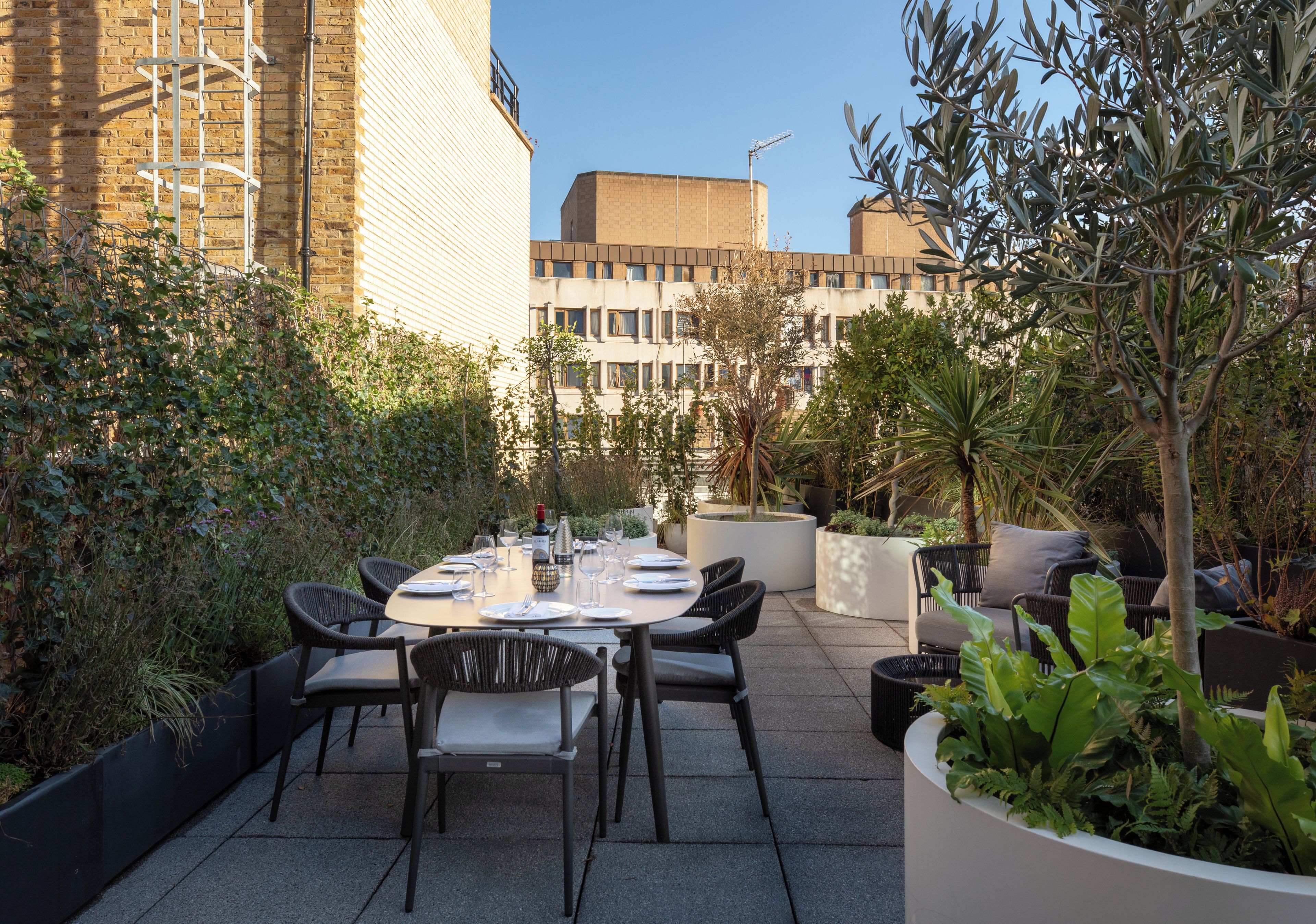 An outdoor dining area with a round table set for four, surrounded by chairs, in a courtyard with buildings in the background.