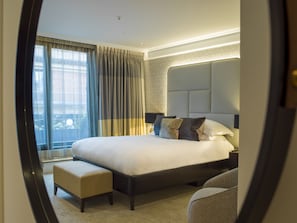 Habitación de hotel moderna con una cama grande, un banco, una mesita de noche y vistas de la ciudad a través de amplias ventanas.