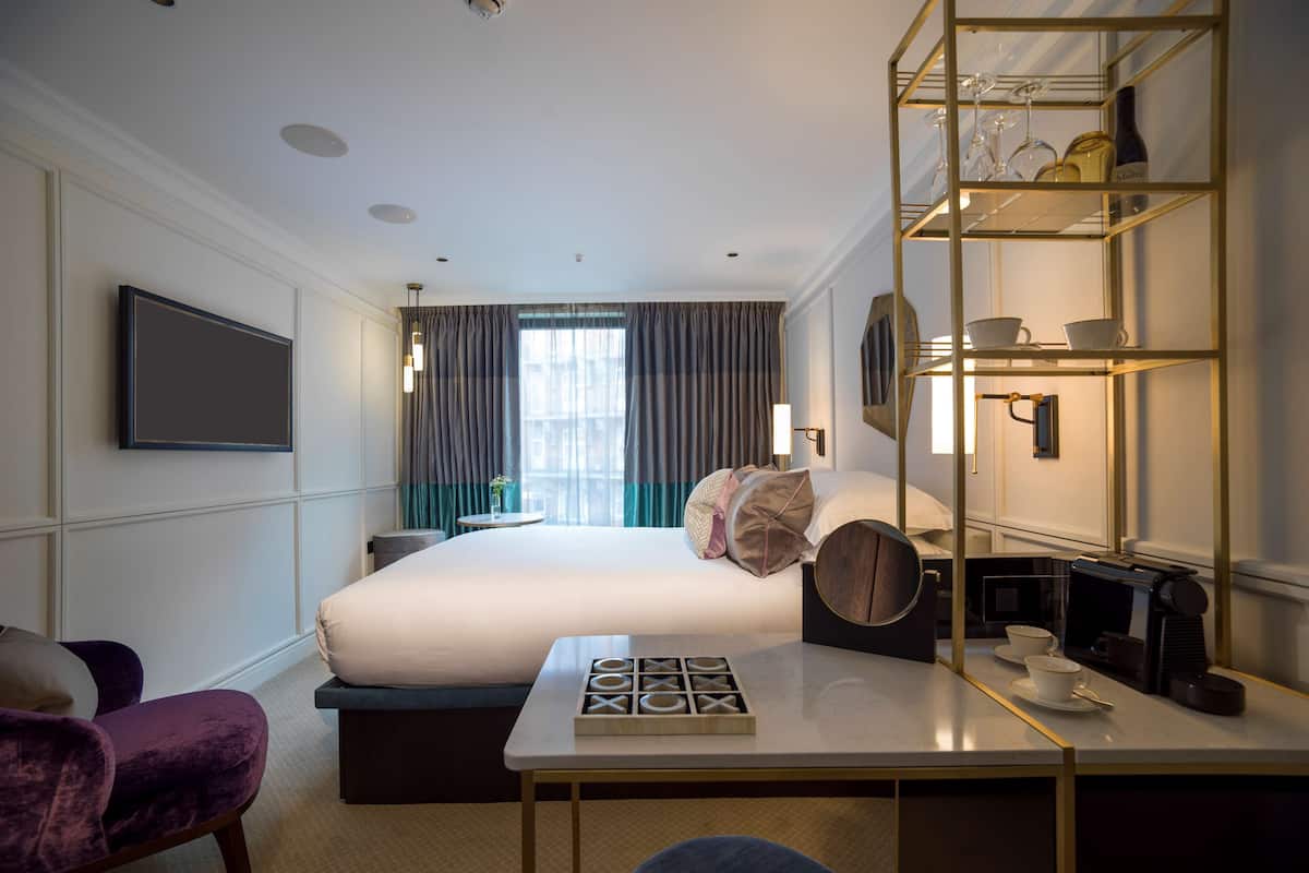 A hotel room with a large bed, a desk with a coffee maker, a TV mounted on the wall, and a shelf with glasses and cups.