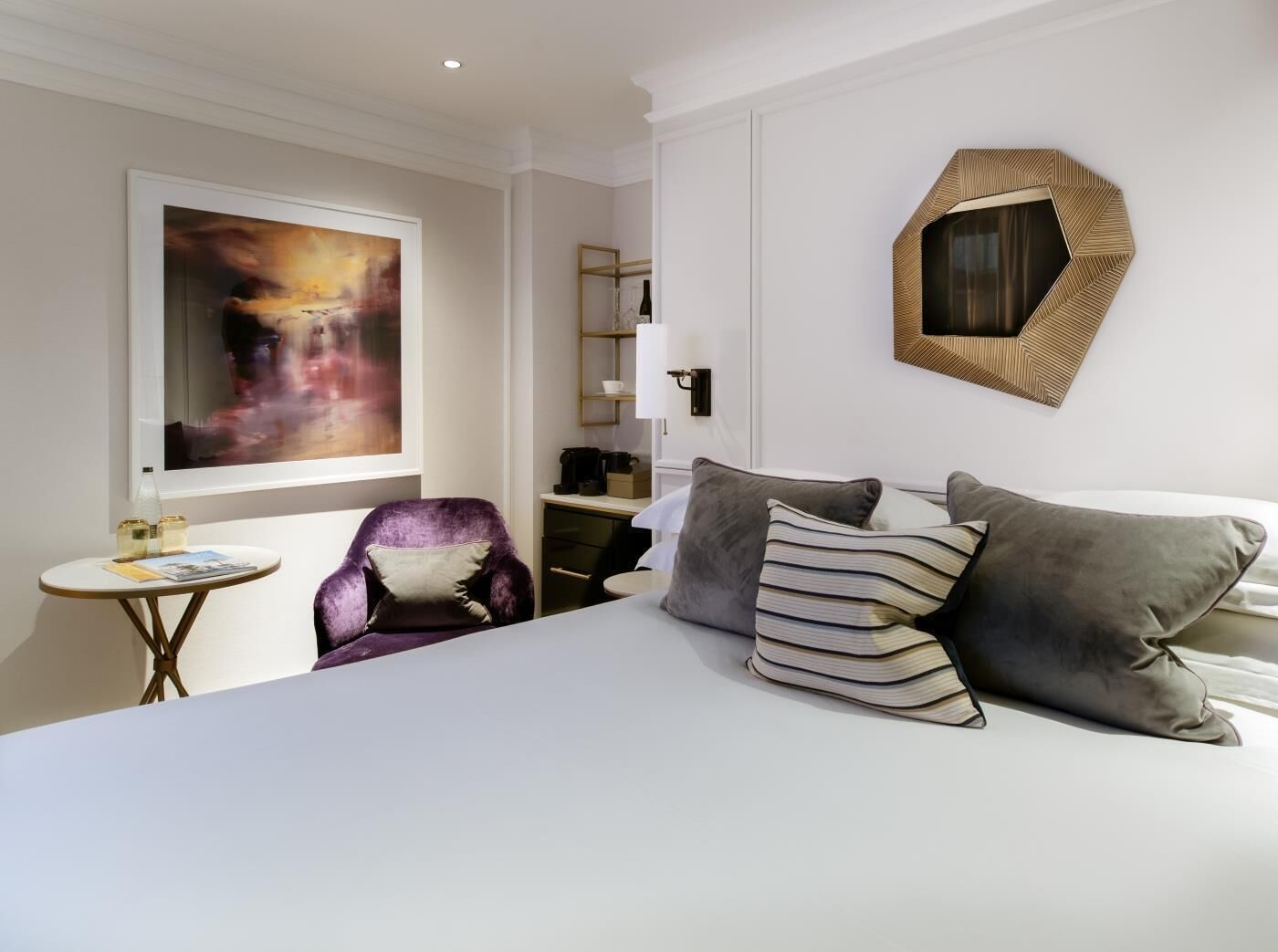 A hotel room with a large bed, a chair, a small table, and a painting on the wall.