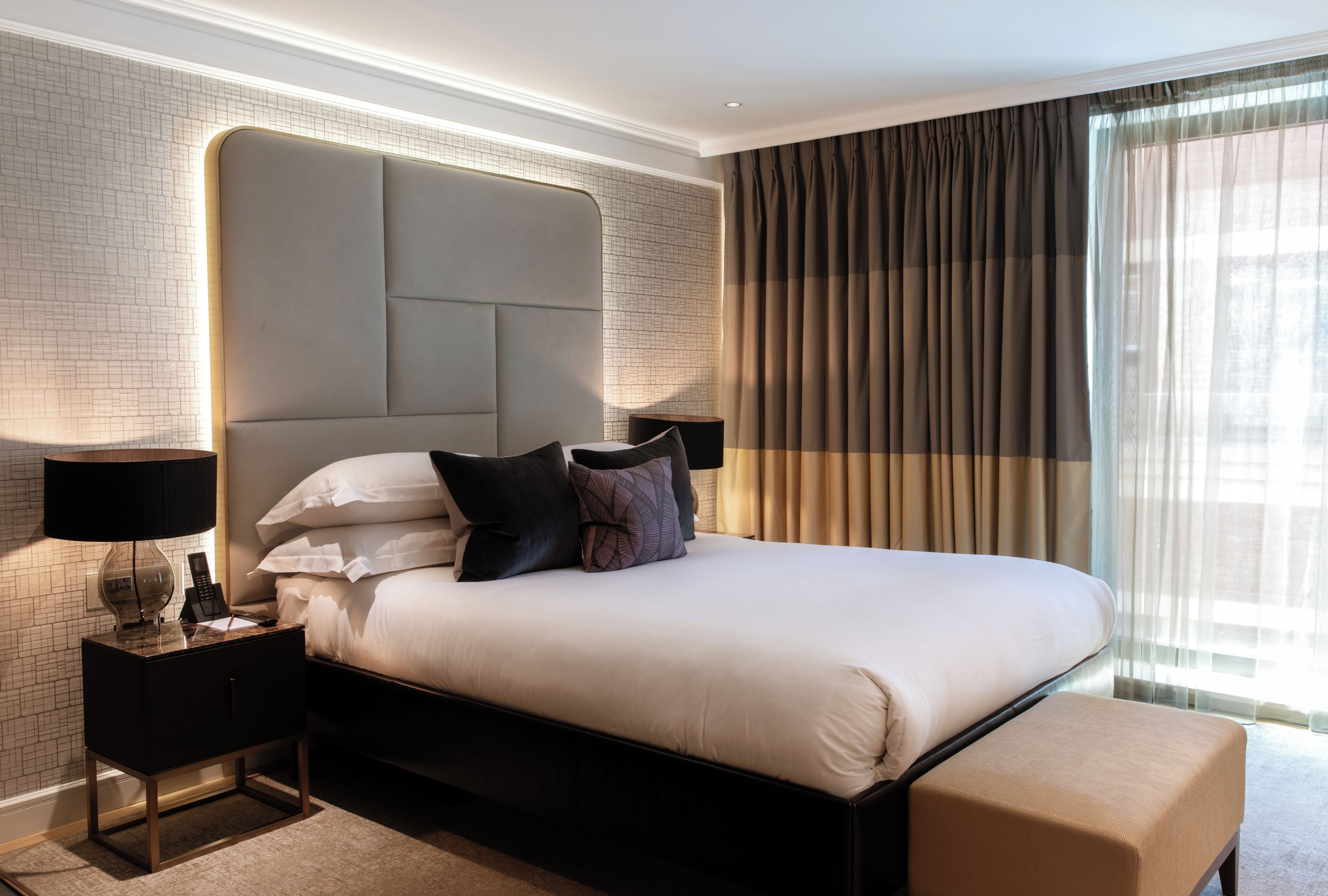 A hotel room with a large bed, a headboard, a bedside table with a lamp, and a window with curtains.