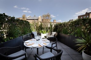 An outdoor patio with a round table set for dining, surrounded by wicker chairs, with a view of buildings in the background.