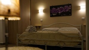 Un lit bien fait, avec un cadre en métal, un tableau représentant des fleurs violettes accroché au mur et une lampe dotée d’un abat-jour beige.