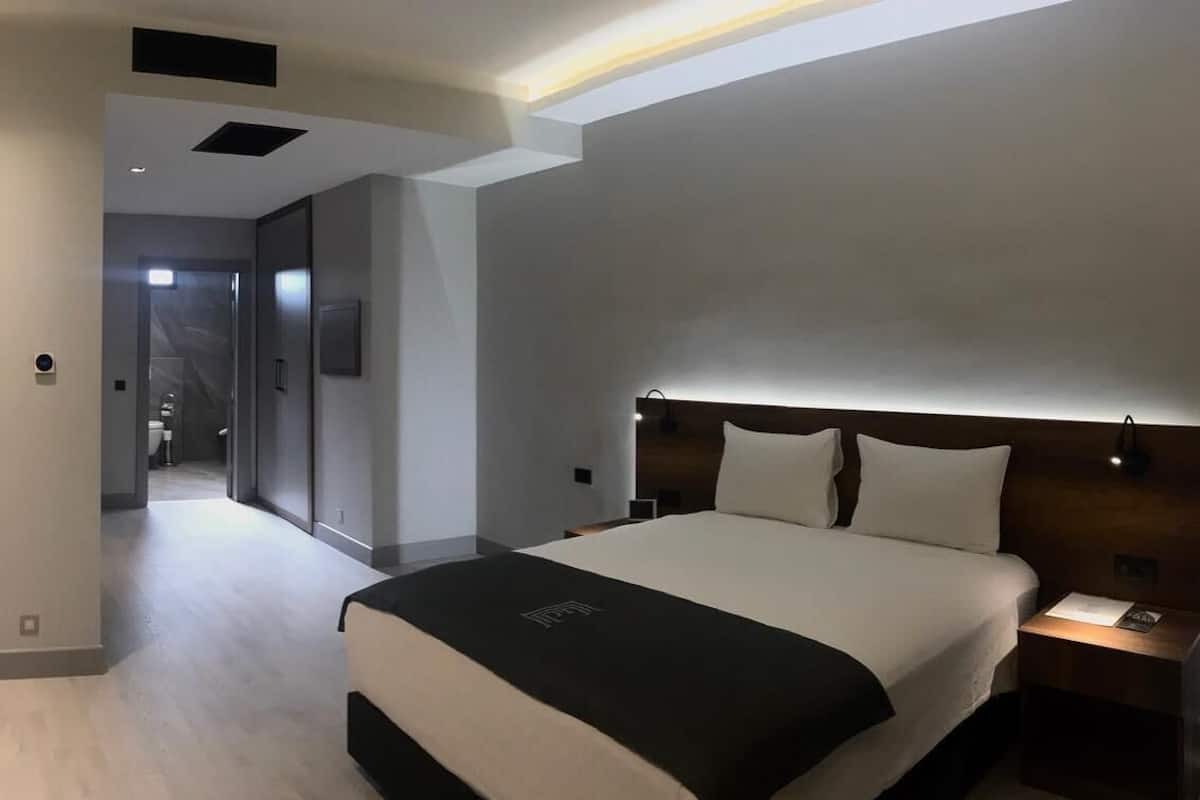 A modern hotel room with a large bed, a desk, and a chair. There is a hallway leading to a bathroom and a door to another room.