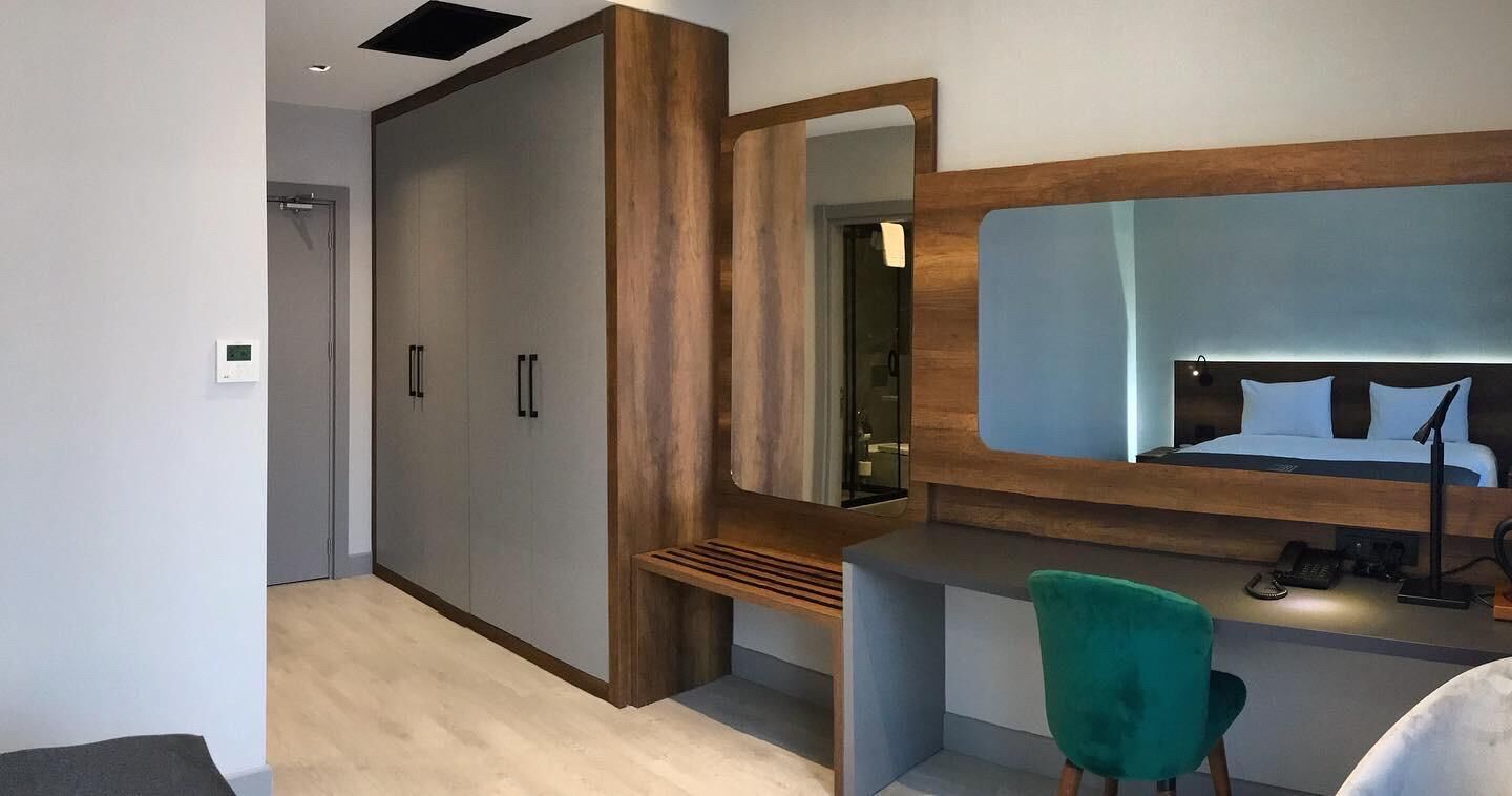 A modern hotel room with a large wardrobe, a vanity with a mirror, a desk, and a green chair.