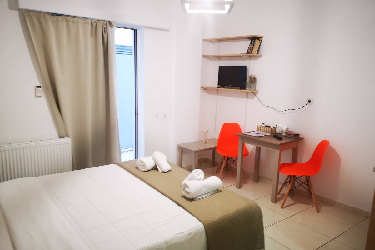 Une chambre d’hôtel avec un lit, un bureau, deux chaises, une télévision et une fenêtre avec des rideaux.