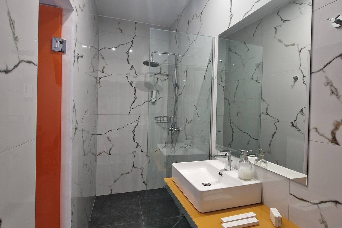 Une salle de bain moderne avec une douche à parois de verre, un lavabo blanc doté d’un plan de toilette en marbre et un grand miroir.