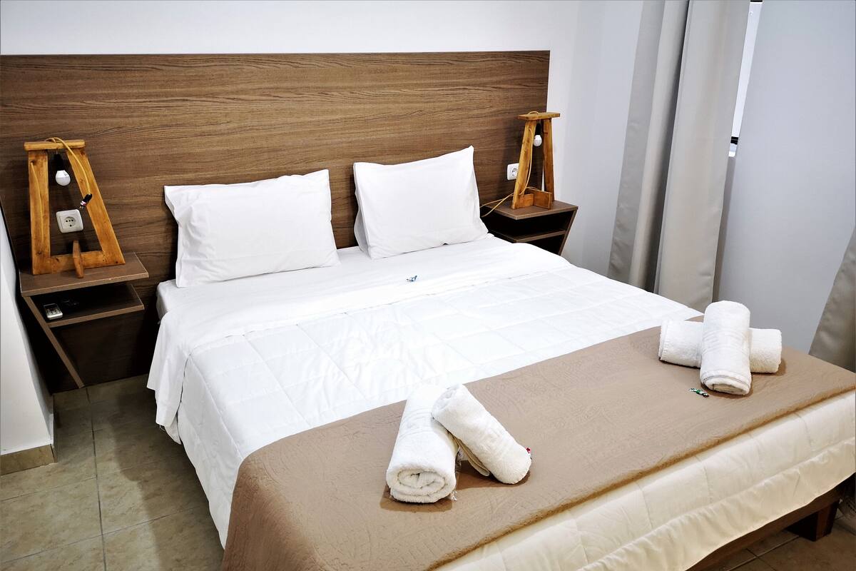 Un lit bien fait, avec du linge de lit blanc, un plaid marron et des serviettes enroulées posées sur le lit. De chaque côté du lit, on trouve des tables de chevet en bois, avec une lampe et une prise électrique.