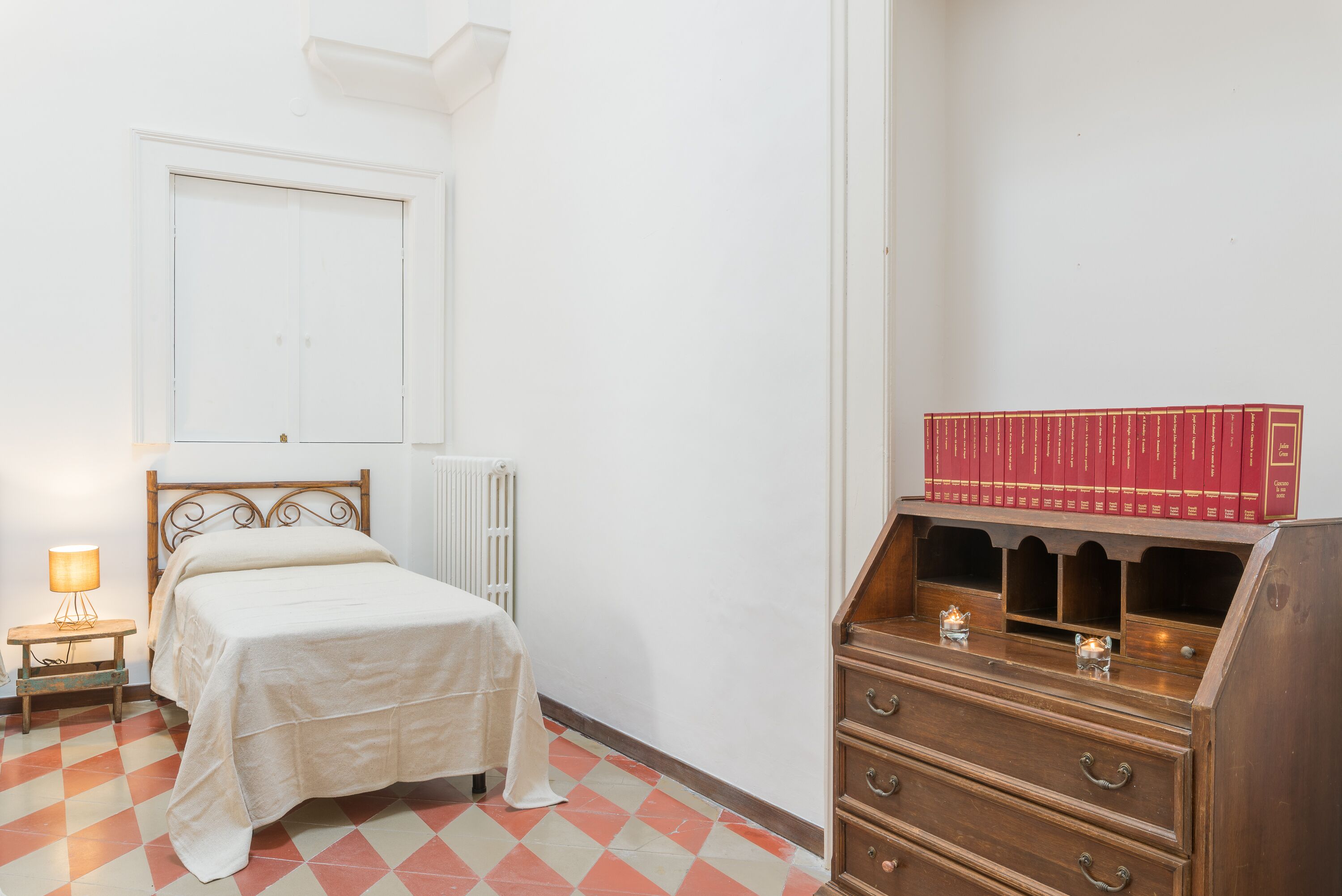 A bedroom with a bed, a nightstand with a lamp, a radiator, and a wooden dresser with books.
